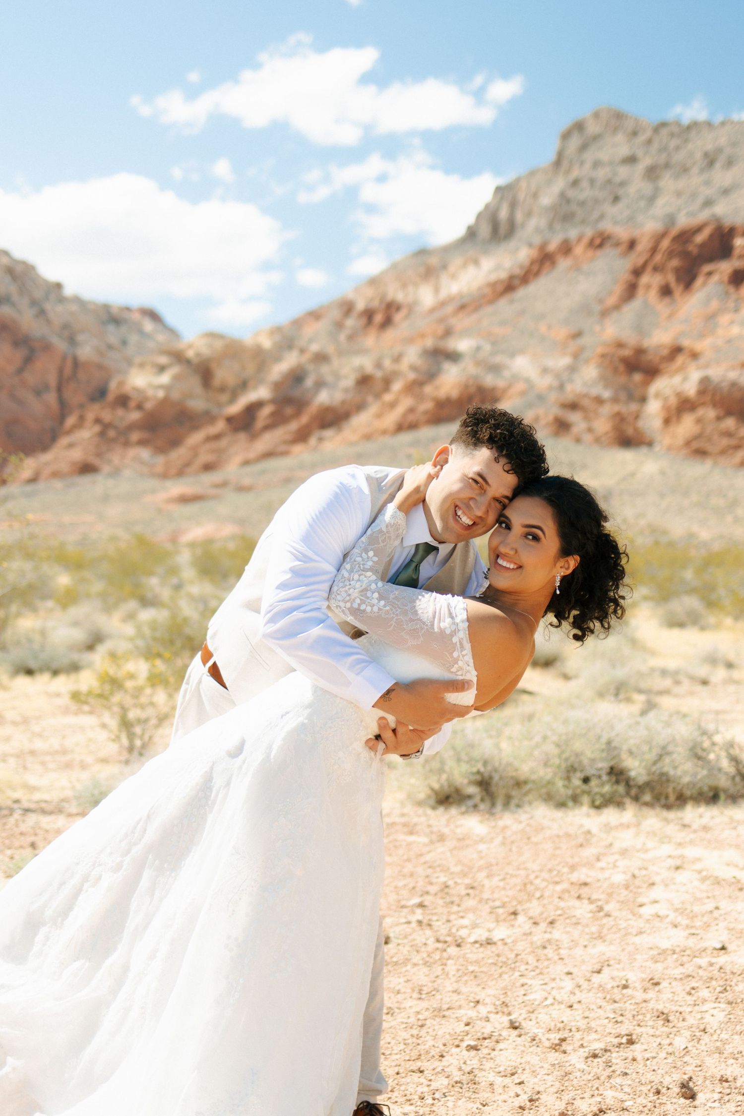 A couple in wedding attire share a romantic dip pose against dramatic red rock formations in a desert landscape.