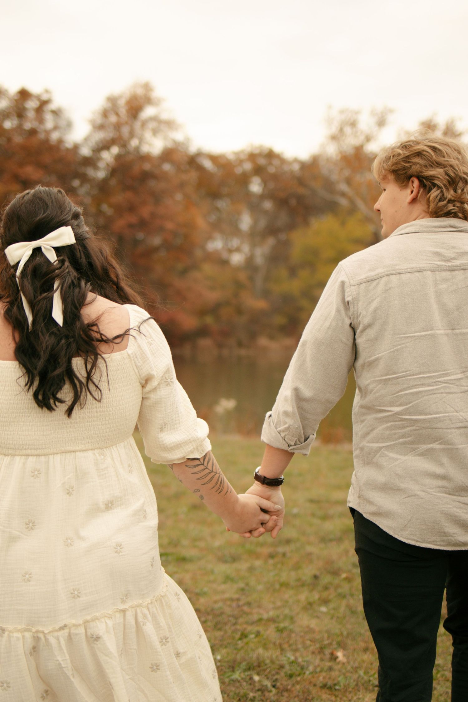 Couple holding hands walking toward autumn lake, woman in white dress with bow, man in gray shirt.