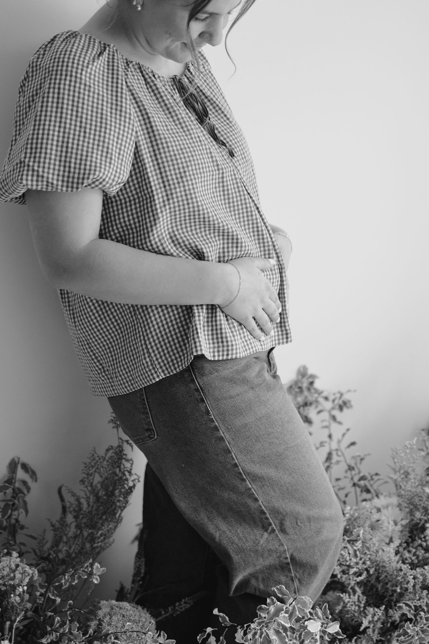 Black and white photo of a pregnant woman in a gingham top and jeans, cradling her baby bump surrounded by plants.