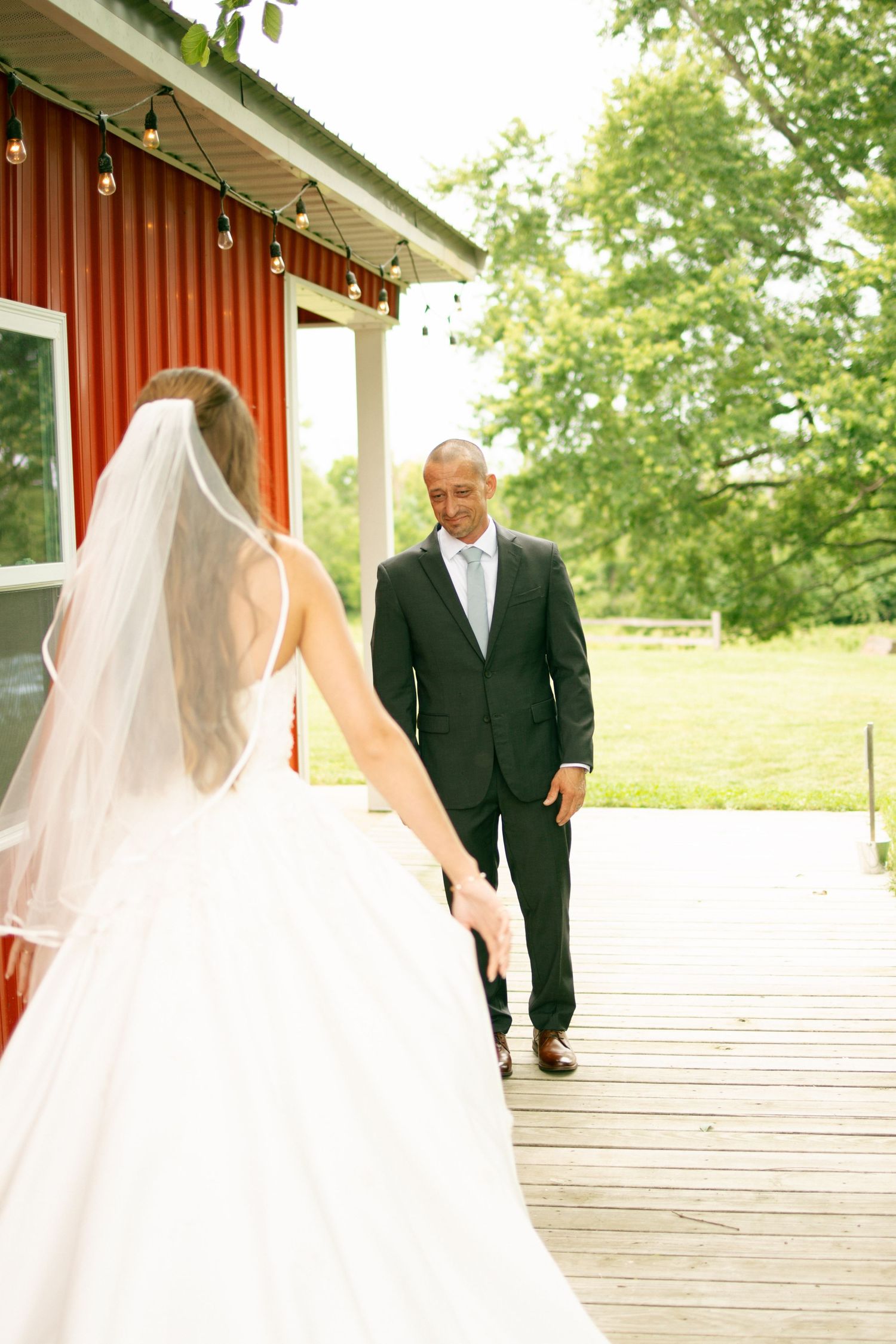 Bride in white gown and veil walks toward groom in black suit on wooden deck outside red barn venue.