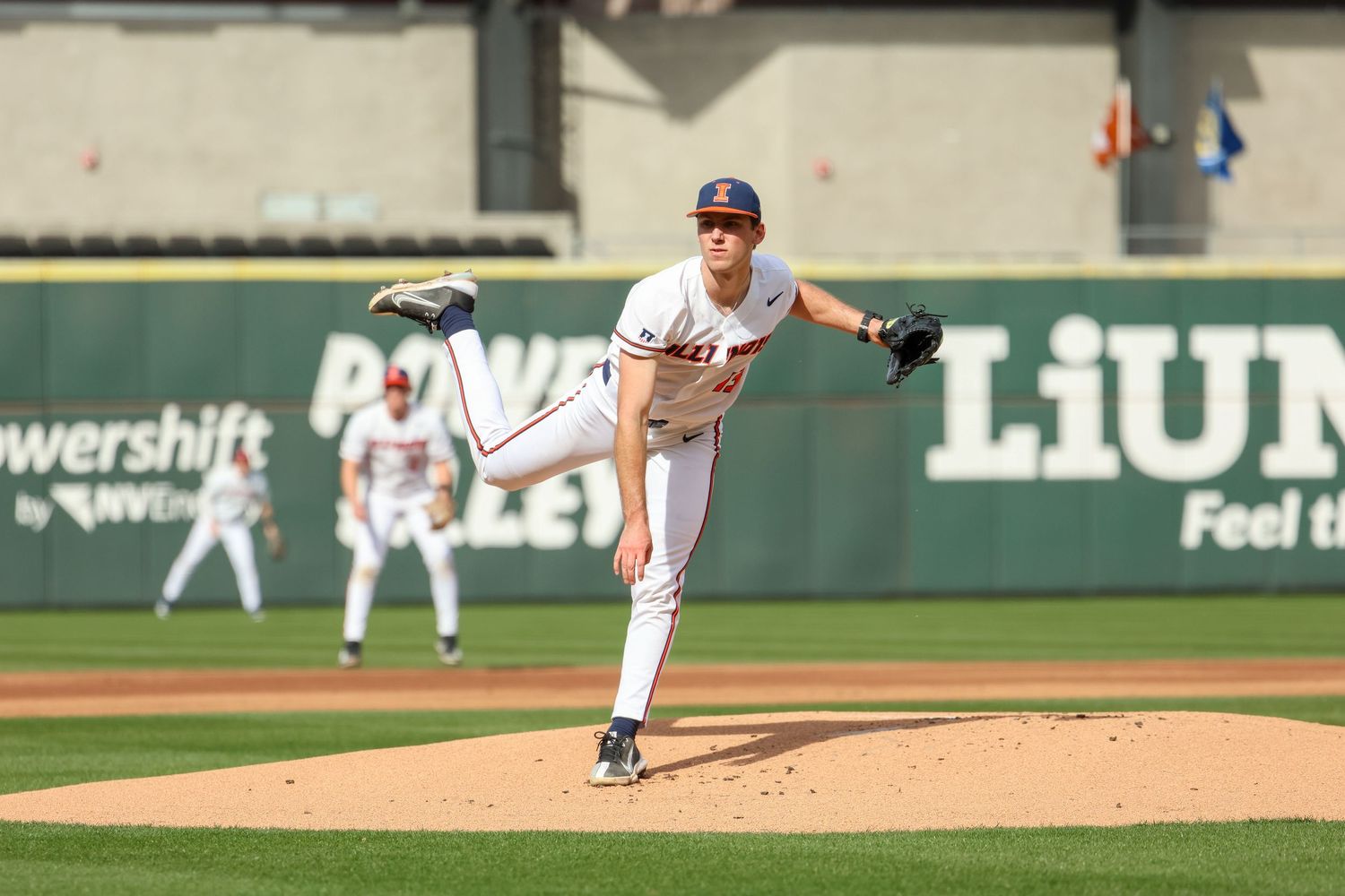 Baseball pitcher in mid-delivery showing full extension during a game at a stadium with advertisements visible.