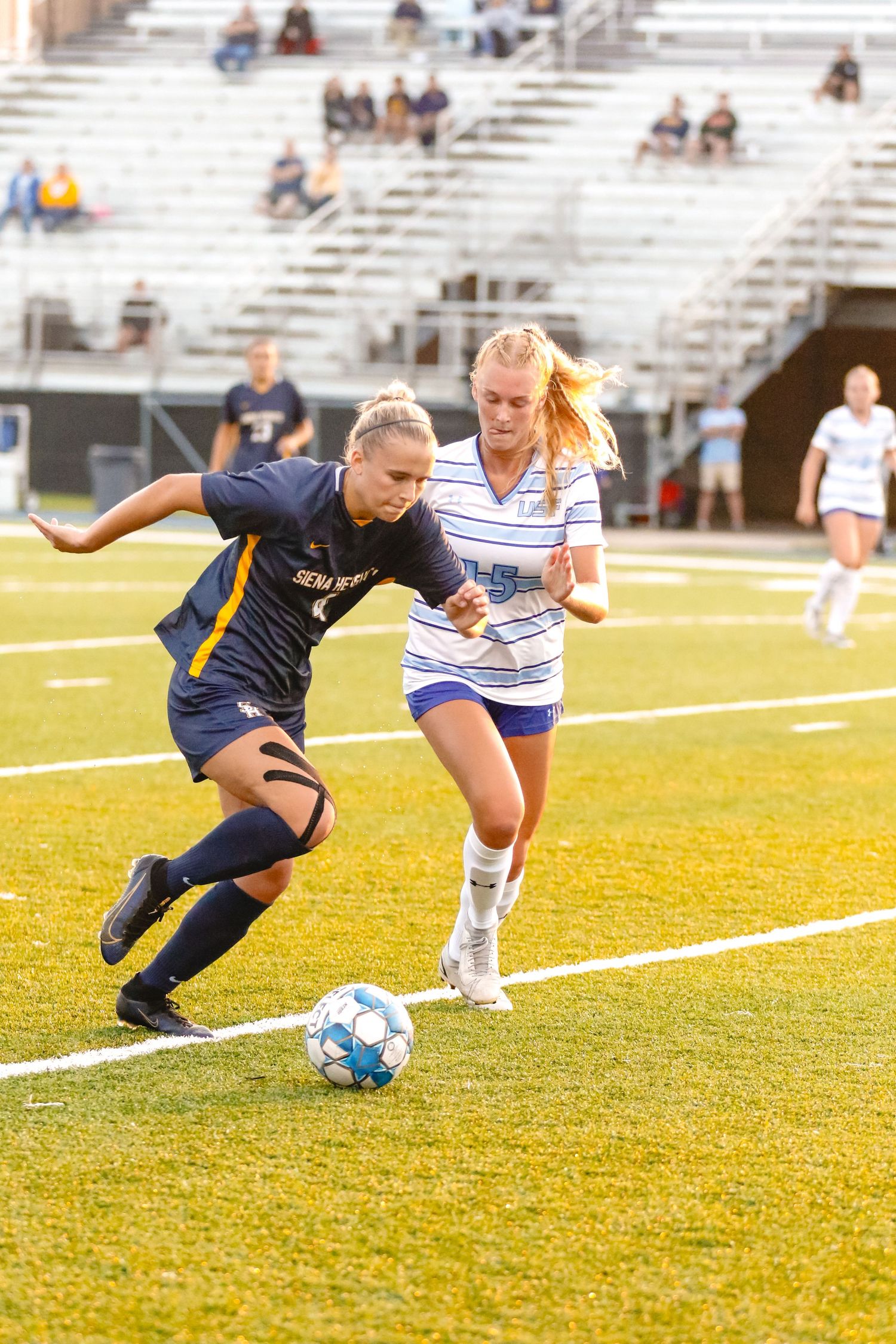 Two soccer players battle for possession of the ball during a competitive match on a sunny grass field.