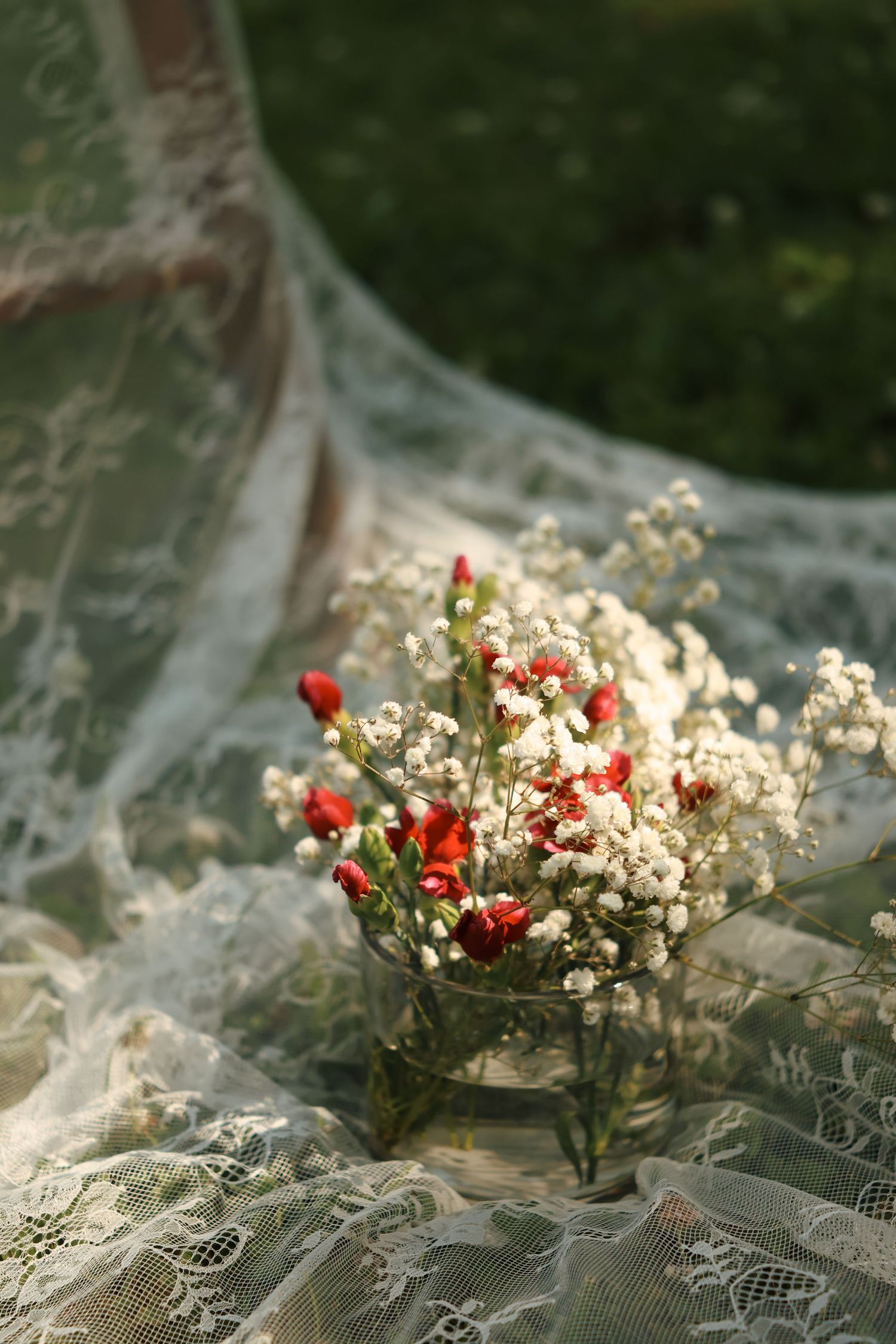 Small glass vase with white baby's breath and red rosebuds on delicate lace fabric outdoors.