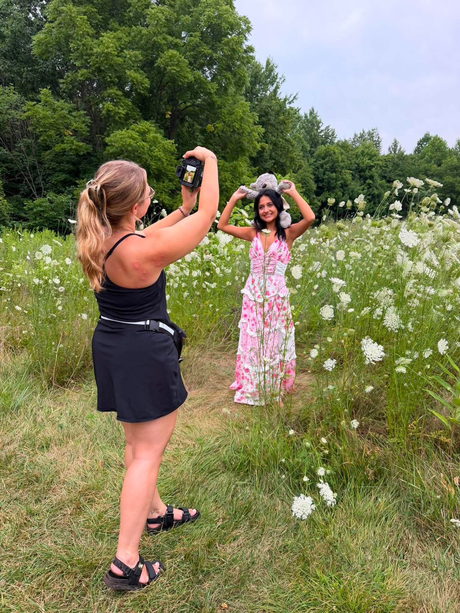 Woman in black dress photographs a woman in floral dress posing in a wildflower meadow surrounded by green trees.