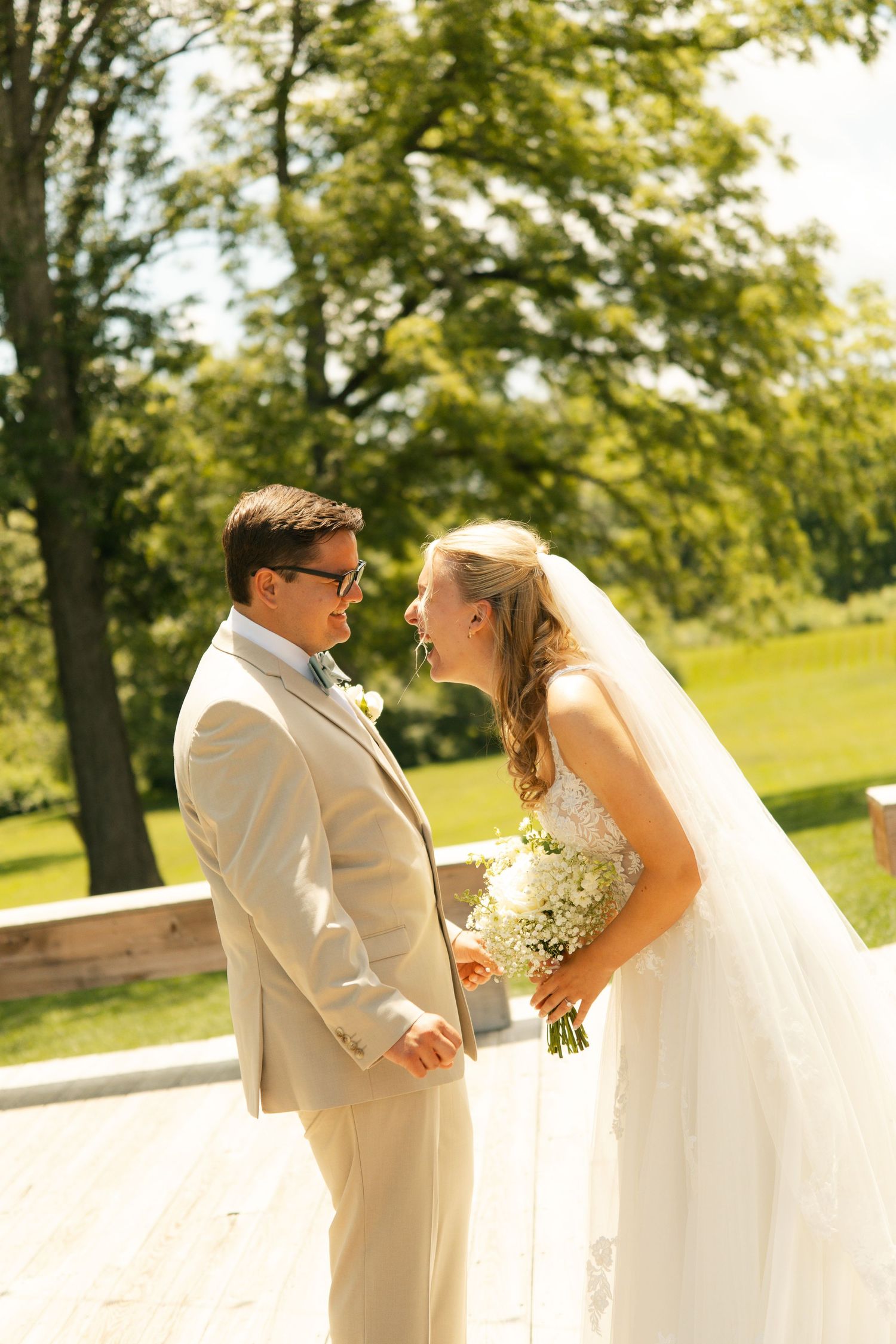 Bride and groom share a joyful moment outdoors, with bride holding white bouquet, groom in tan suit, surrounded by lush greenery.