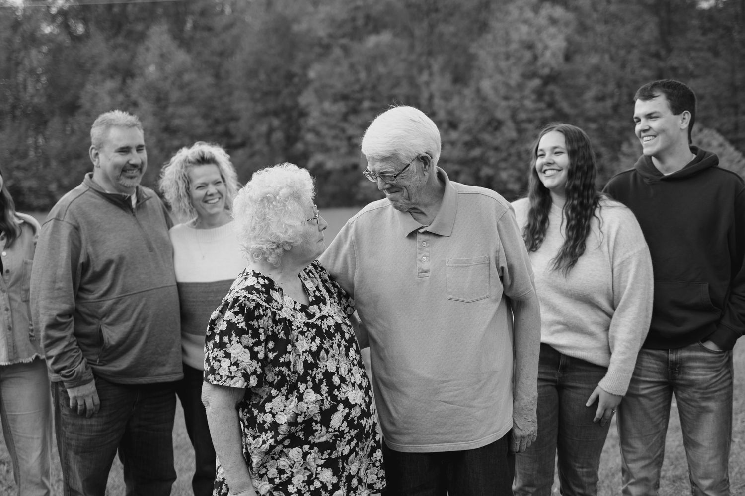 Black and white family photo of elderly couple sharing tender moment surrounded by smiling adult children outdoors.