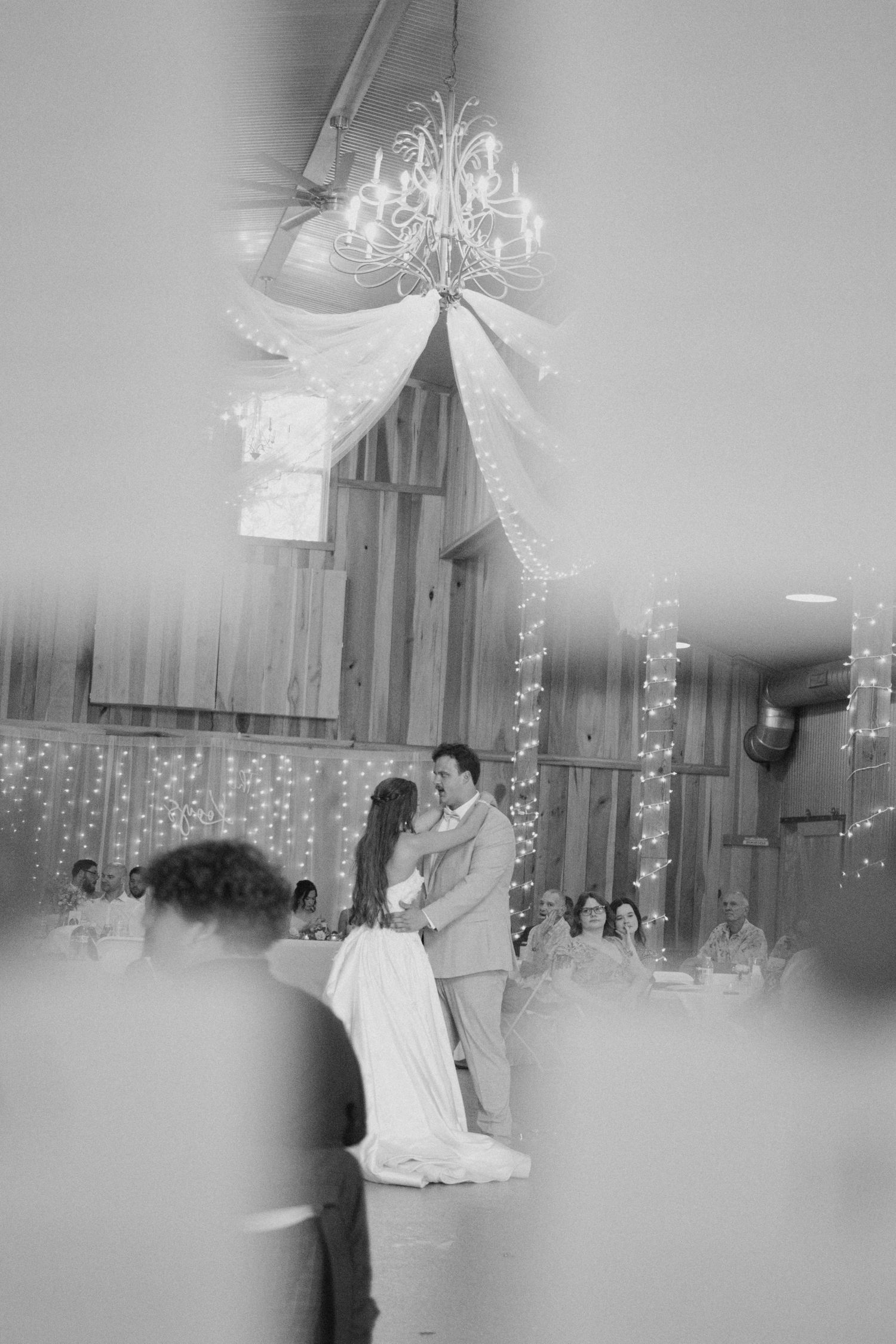 Black and white photo of couple sharing first dance in rustic barn with chandelier and fairy lights.