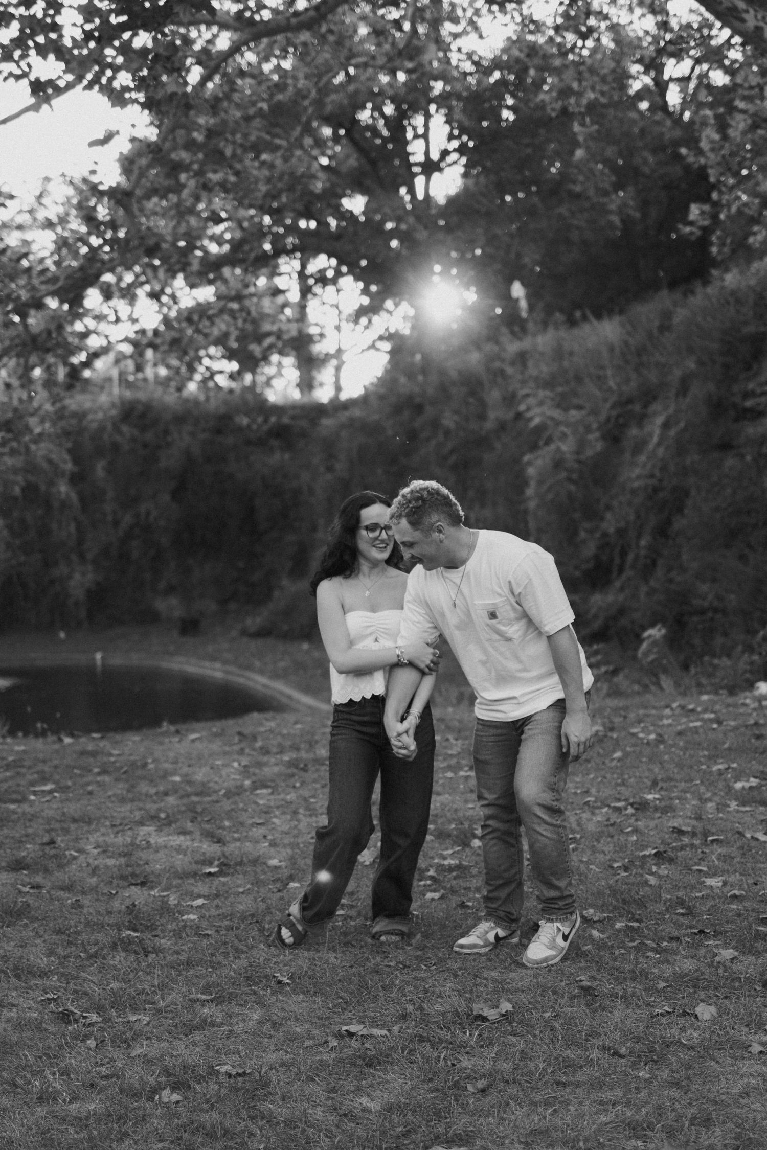 Black and white photo of a couple holding hands and laughing together in a lush park setting at sunset.