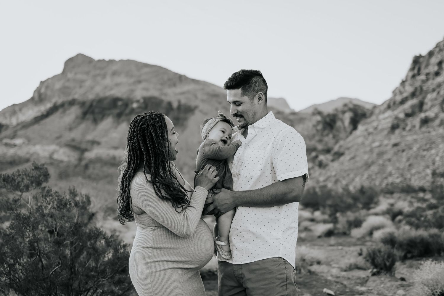 Black and white photo of a happy family with a pregnant woman and man holding a toddler outdoors near rocky mountains.