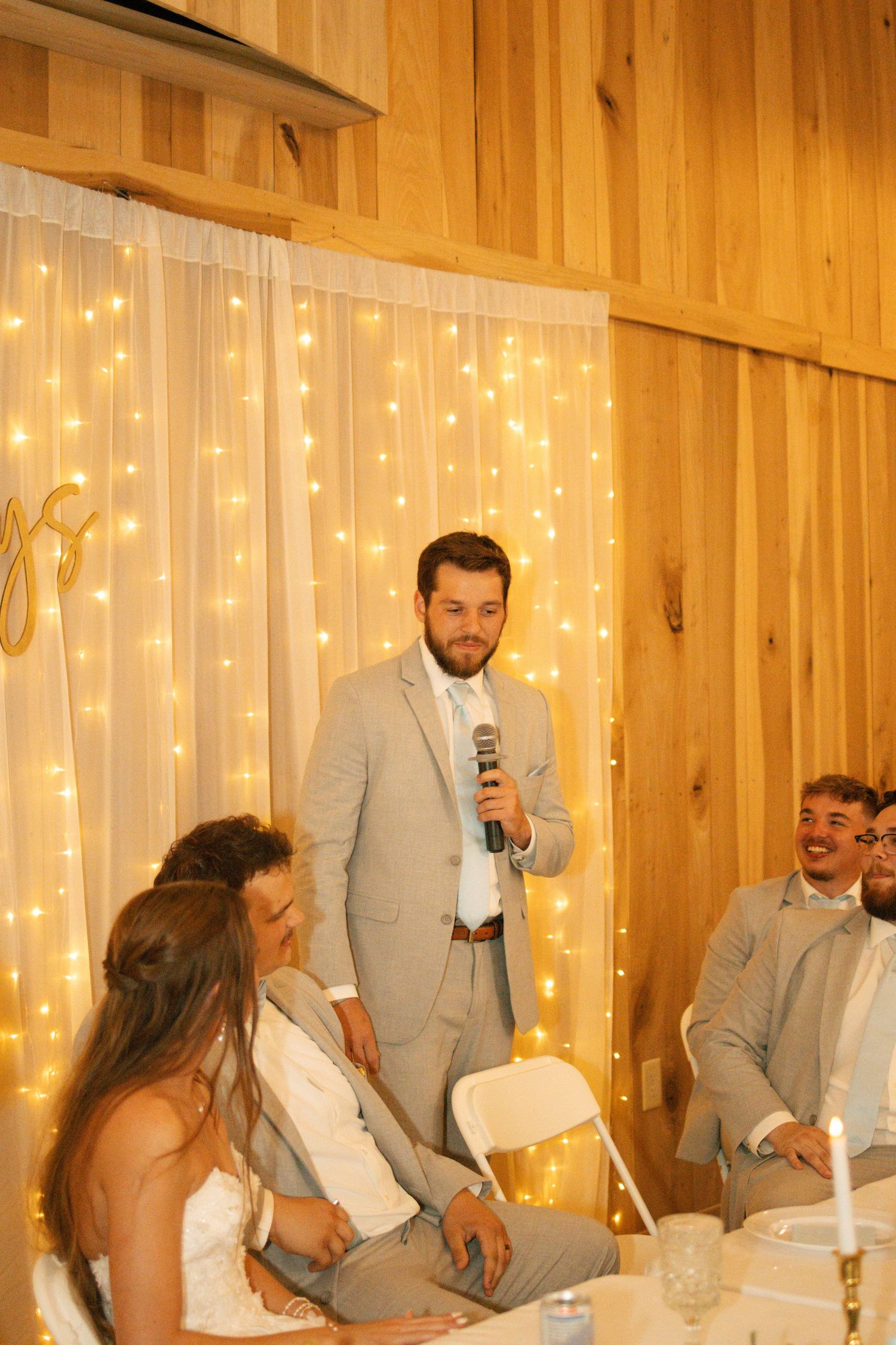 Man in gray suit giving wedding toast with microphone, bride and groom seated at reception table with fairy lights backdrop.