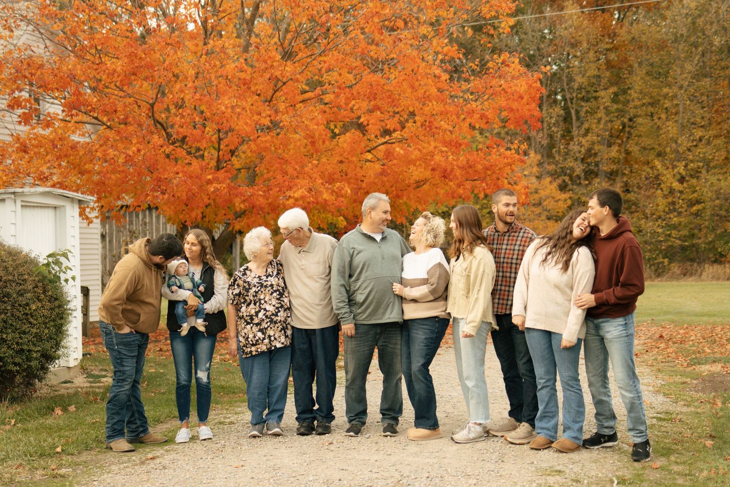 Multi-generational family gathered outdoors in autumn, smiling together under vibrant orange fall foliage trees.