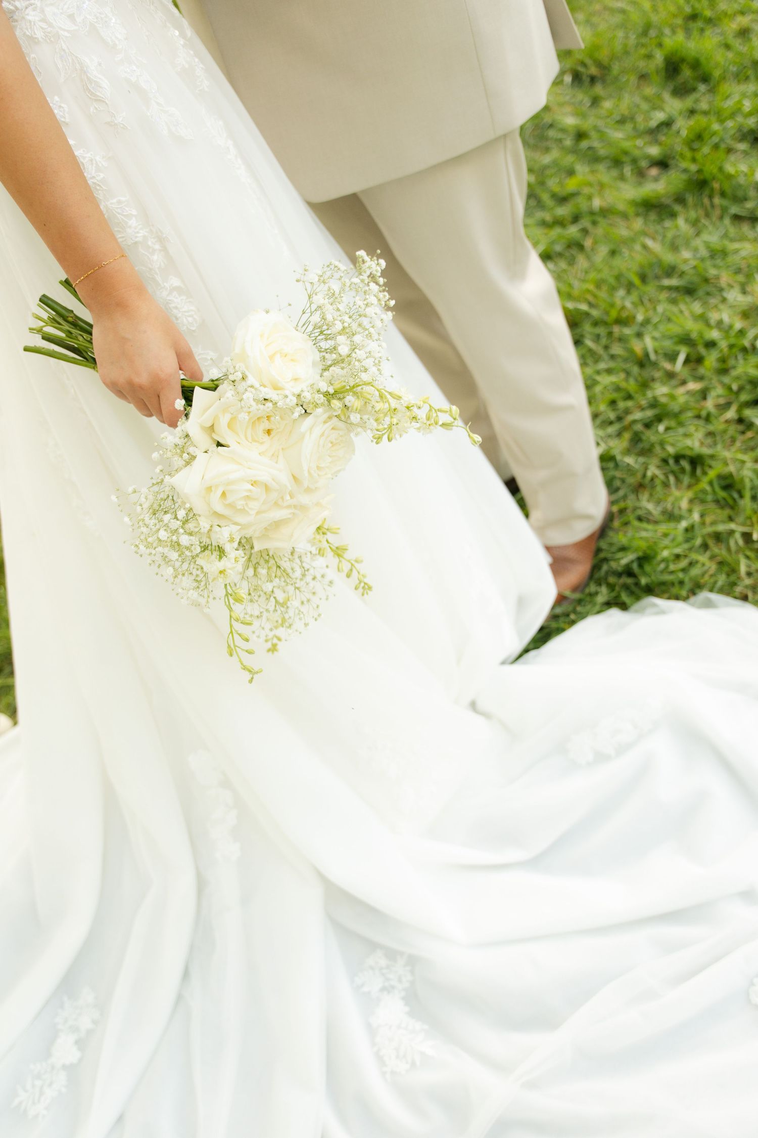 Bride in white lace gown holding elegant white rose bouquet with baby's breath, groom in beige suit on green grass.