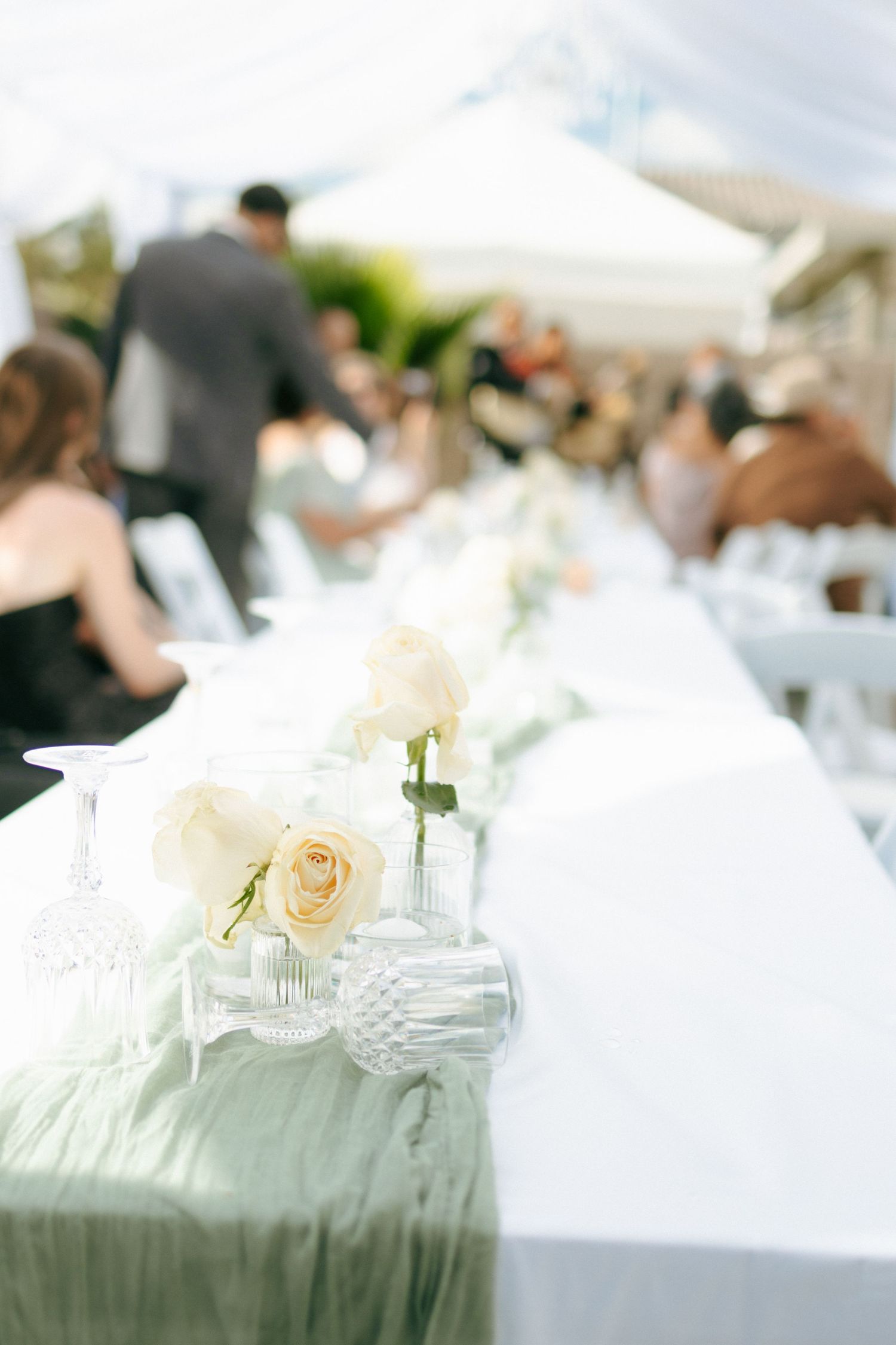 White roses and elegant glassware decorate a wedding reception table with a sage green runner.