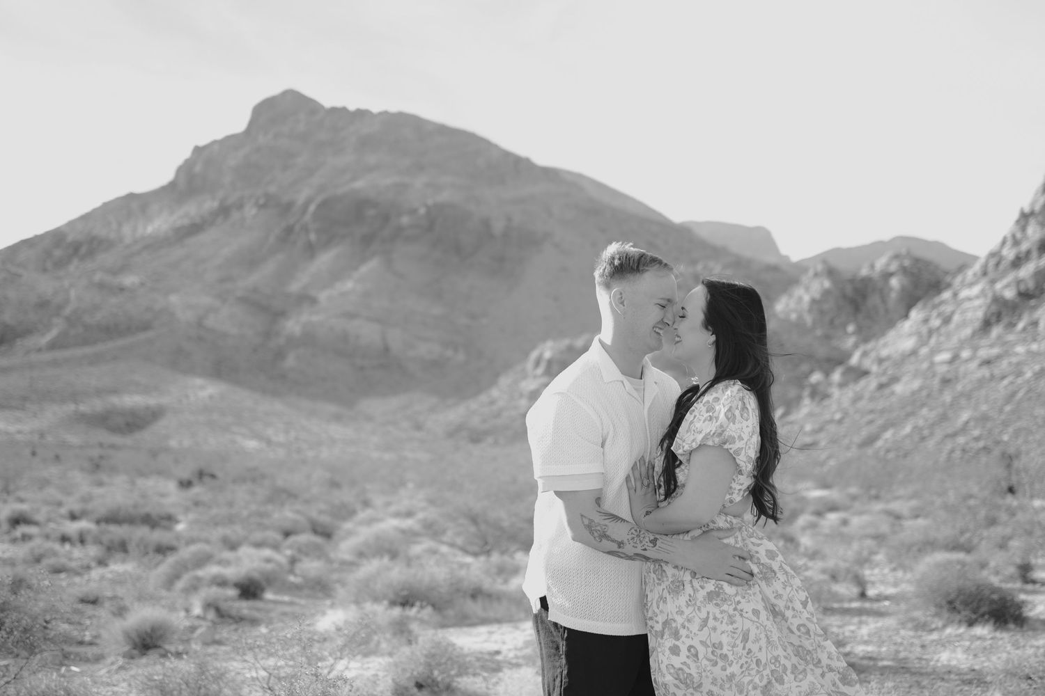A couple shares a romantic moment in front of a desert mountain landscape in black and white.