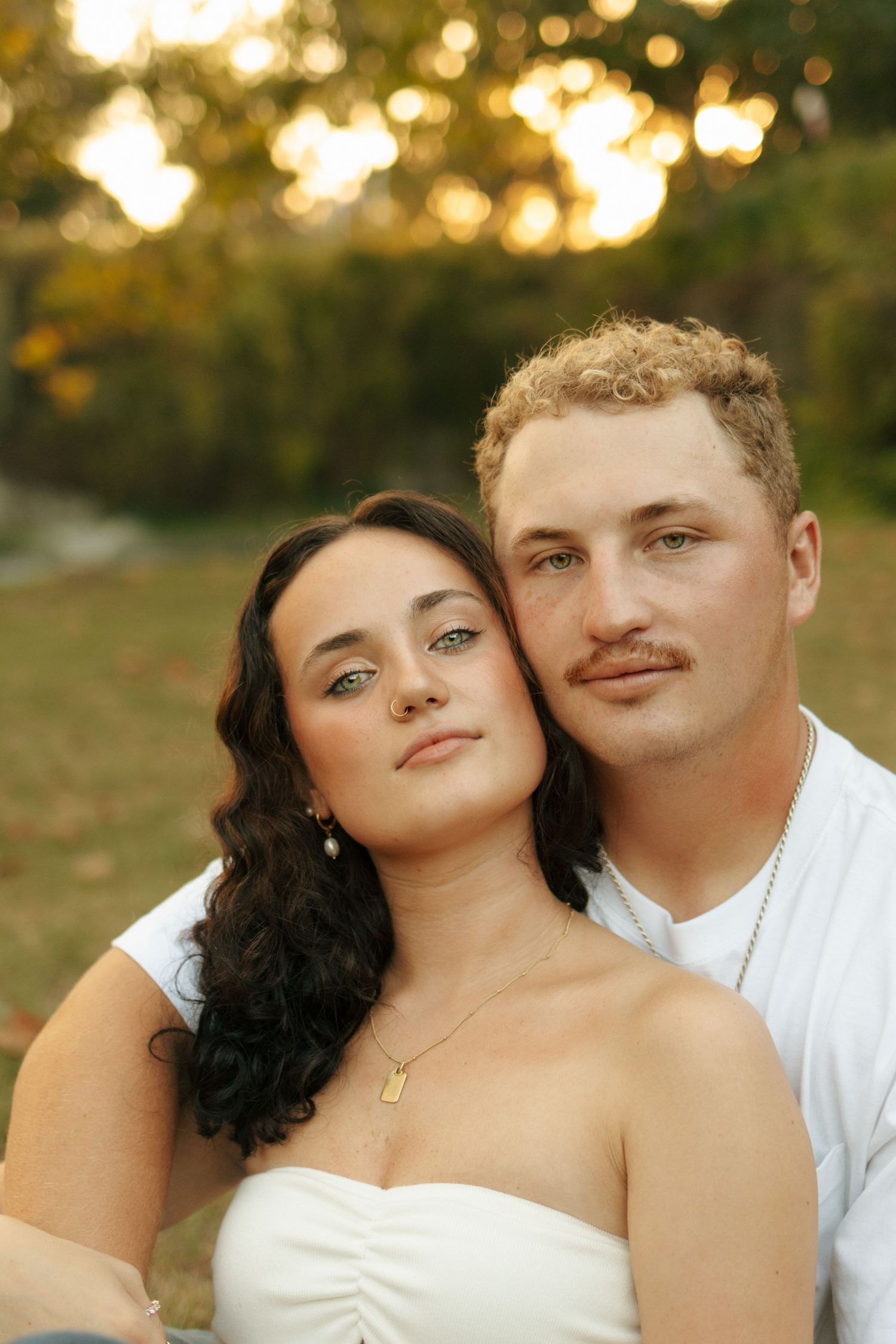 A young couple poses outdoors at golden hour, woman in white dress leaning on mans shoulder among autumn trees.