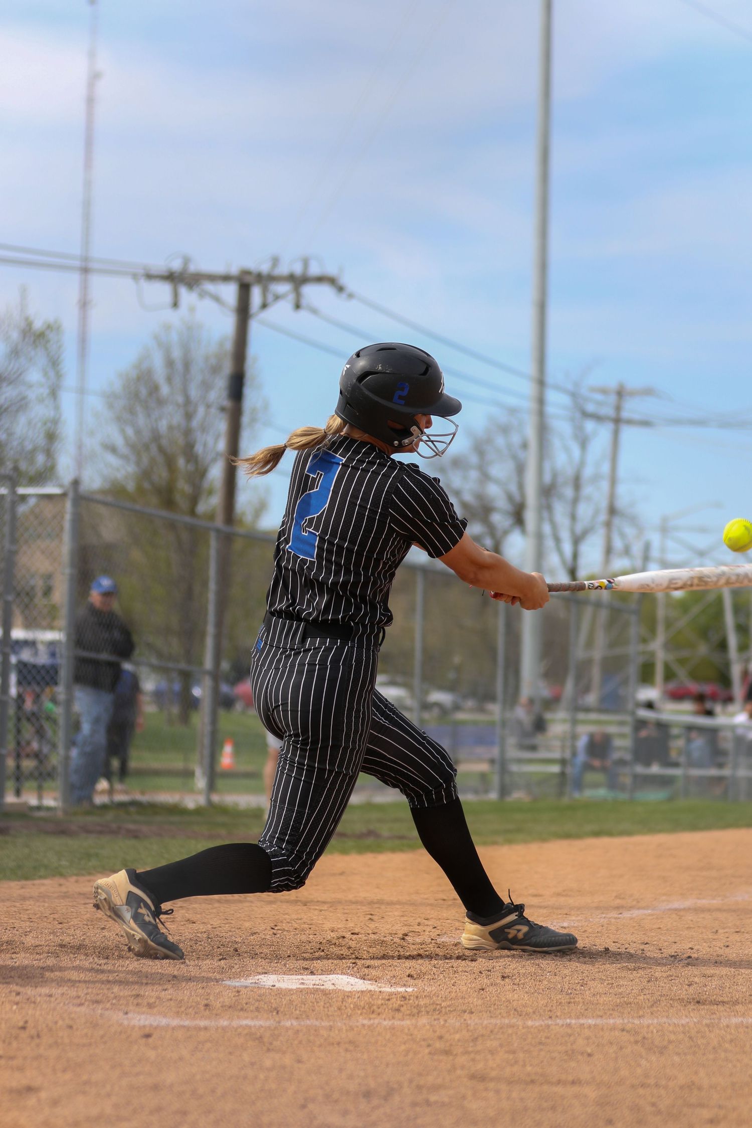 Softball player in pinstriped uniform and helmet swings bat at home plate during game on dirt diamond.