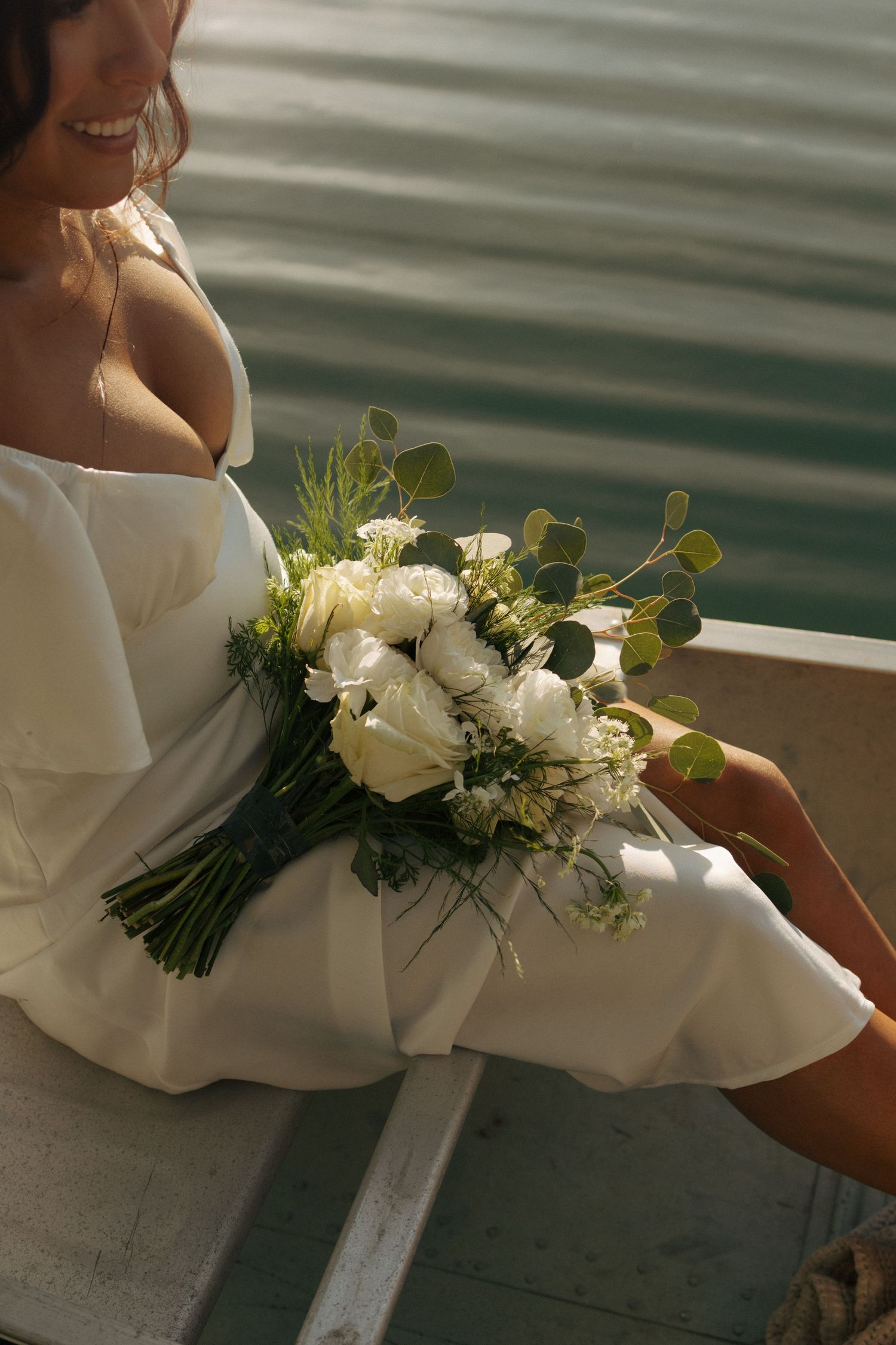 Smiling bride in white off-shoulder dress holding elegant white rose and eucalyptus bouquet while seated outdoors.