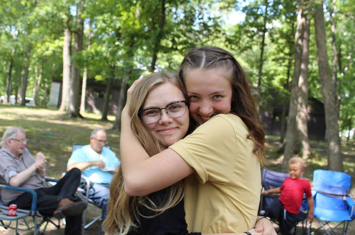 Two young girls share a warm hug outdoors at a wooded park gathering, smiling joyfully with friends seated nearby.
