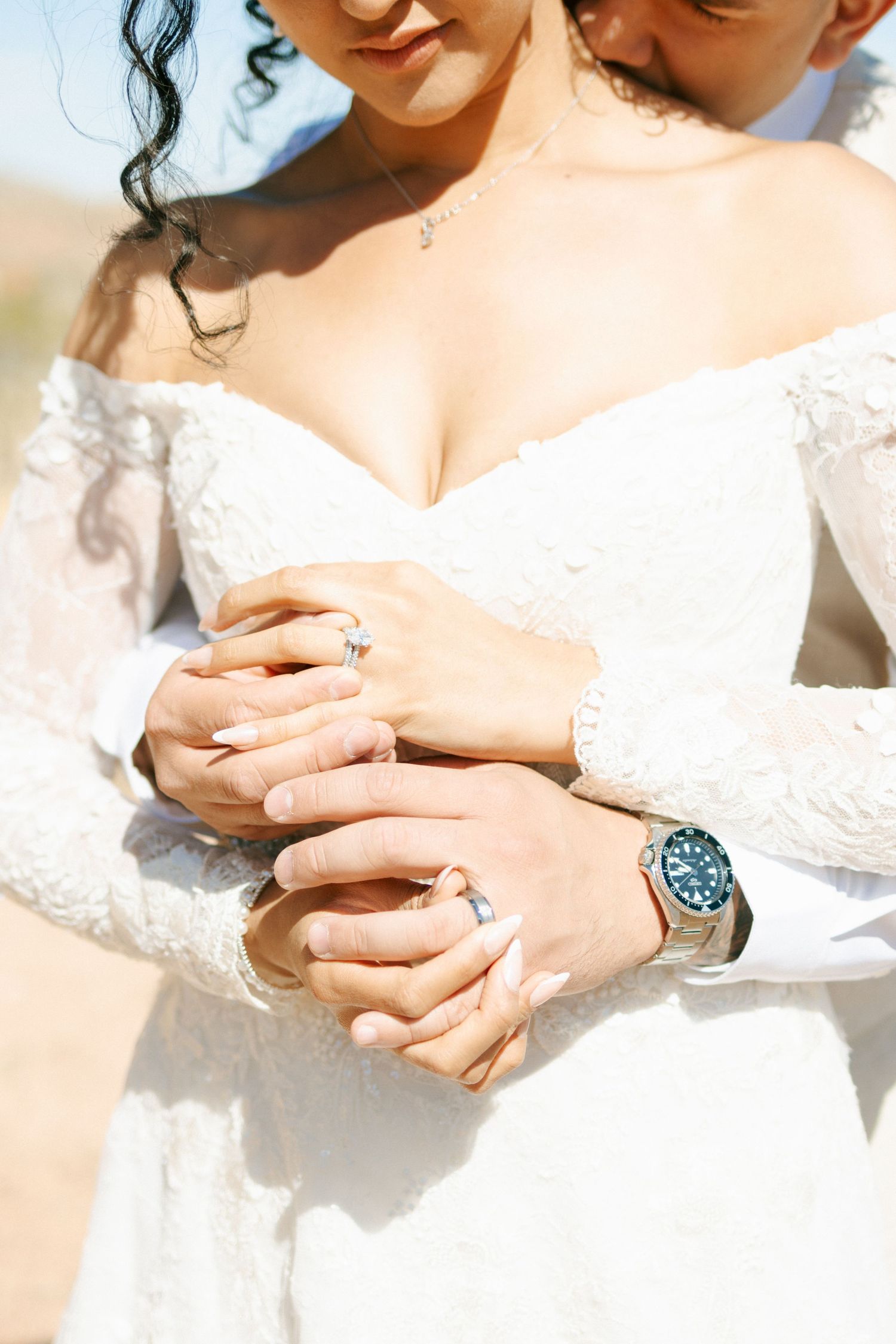 Close up of hands showing wedding rings and lace white dress sleeves with a blue watch visible in sunlight.