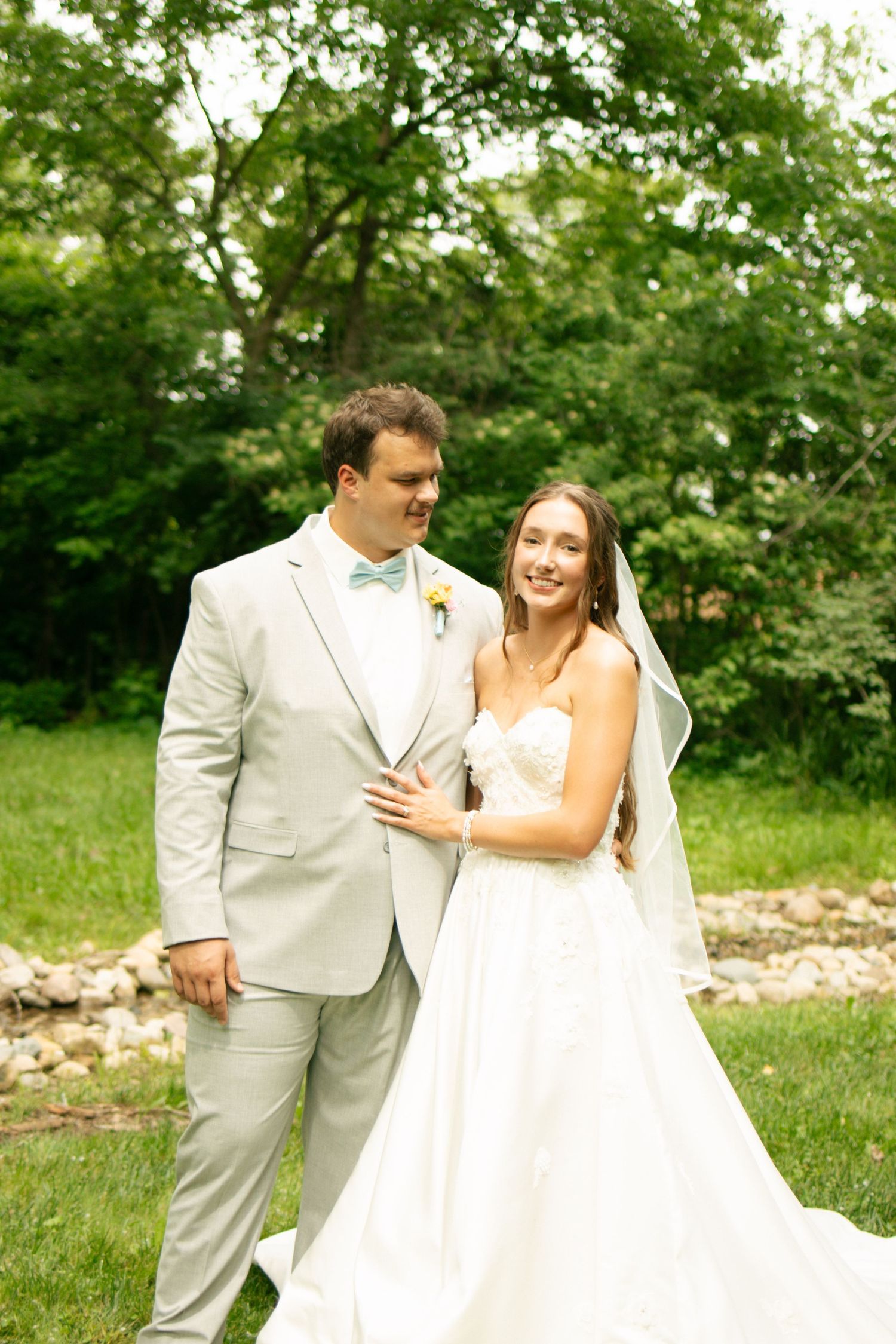 Bride and groom share a joyful moment outdoors, she in a white gown with veil, he in a light gray suit with bow tie.
