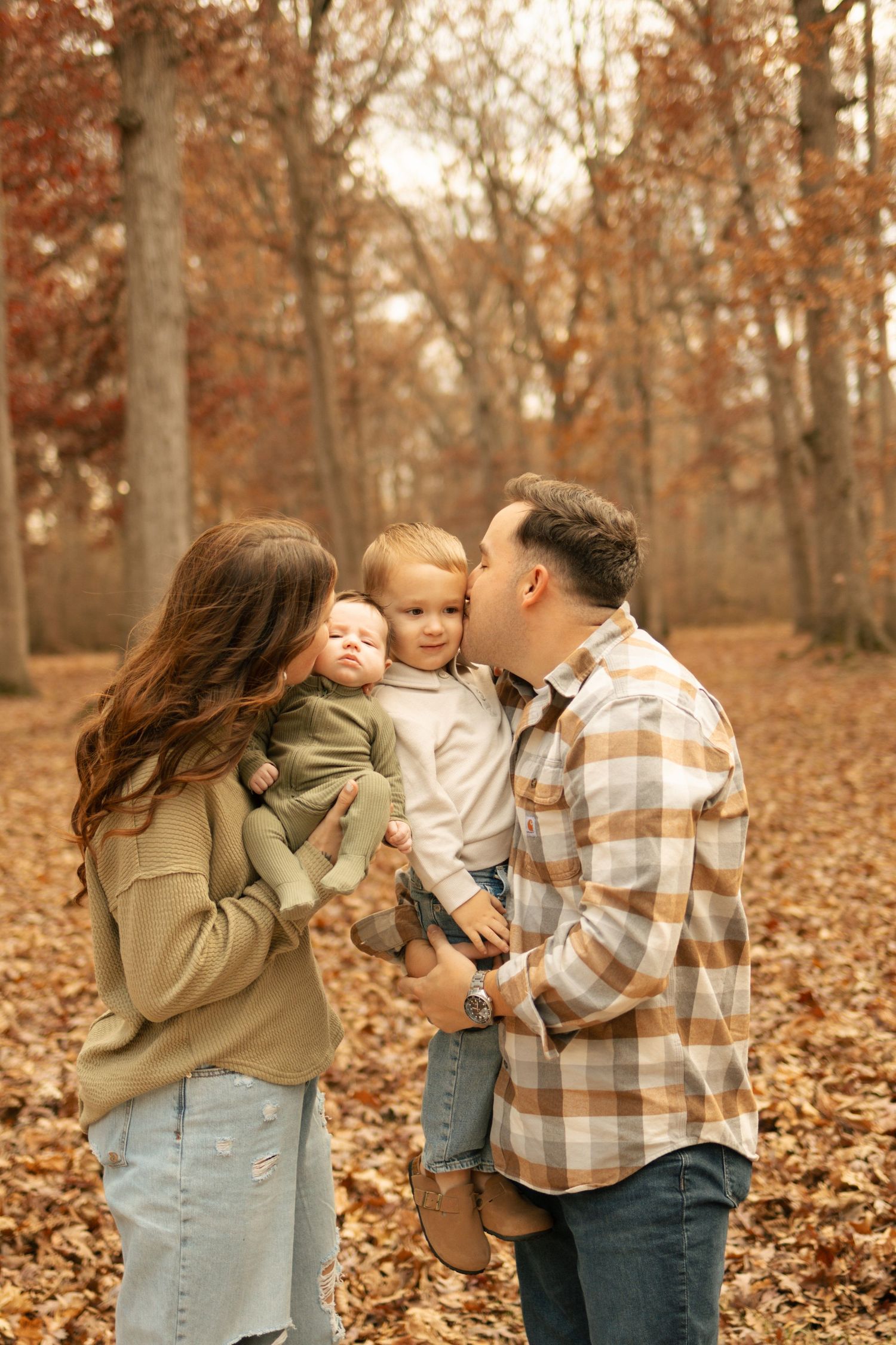 Family of four enjoying an autumn forest walk, parents kissing toddler while holding newborn baby among fall leaves.
