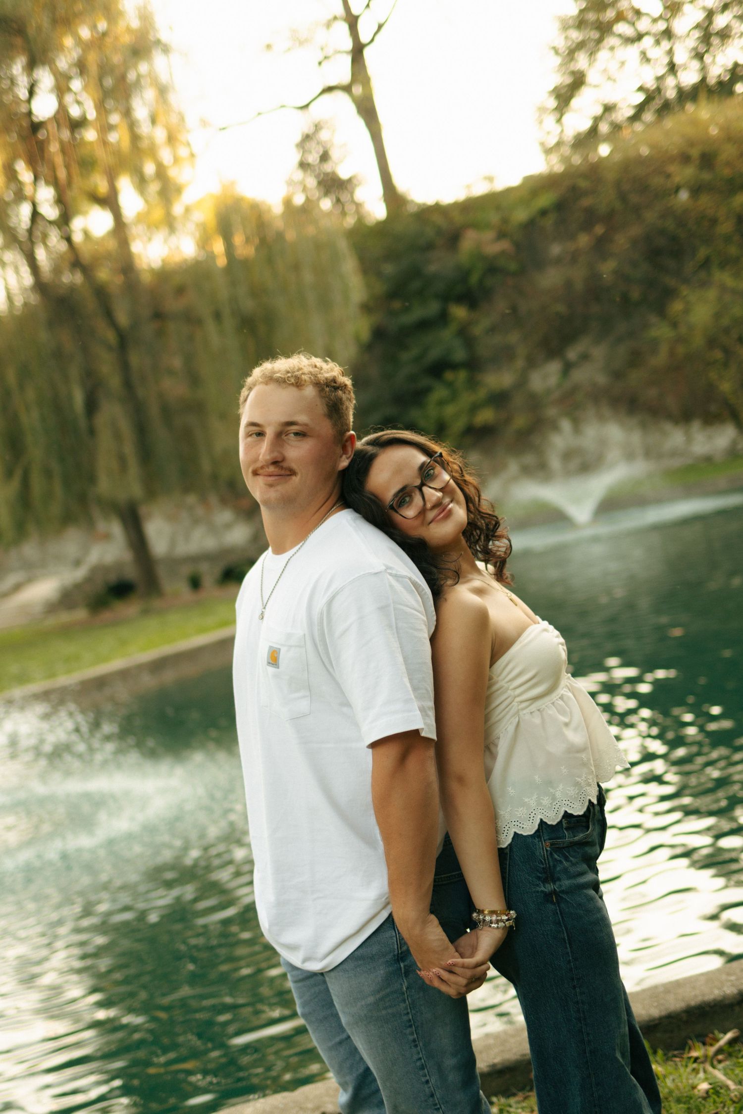 A young couple poses back-to-back near a pond with a fountain, smiling in casual attire during golden hour.