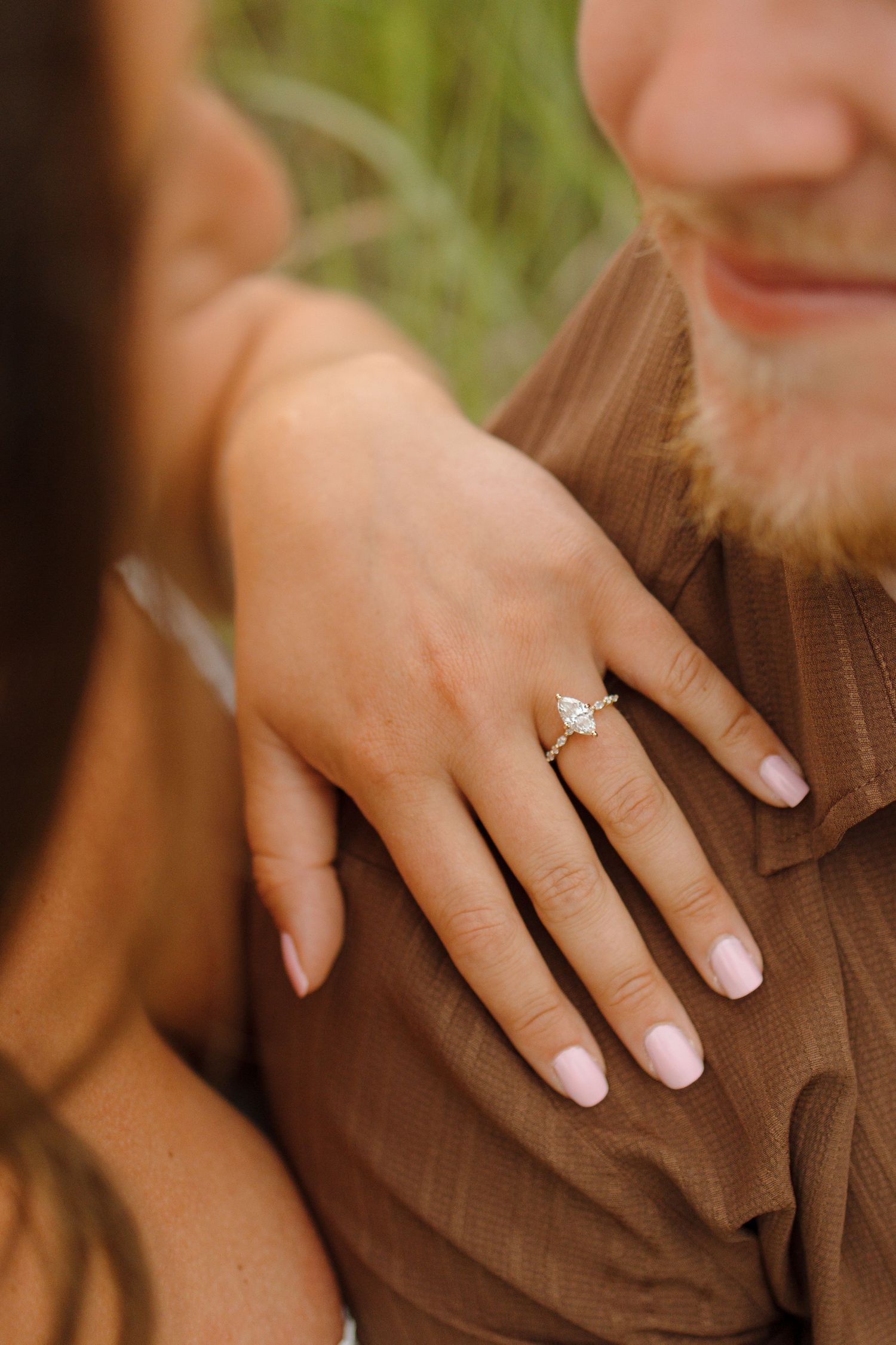 Close-up of a woman's hand displaying a marquise diamond engagement ring resting on a man's brown shirt.