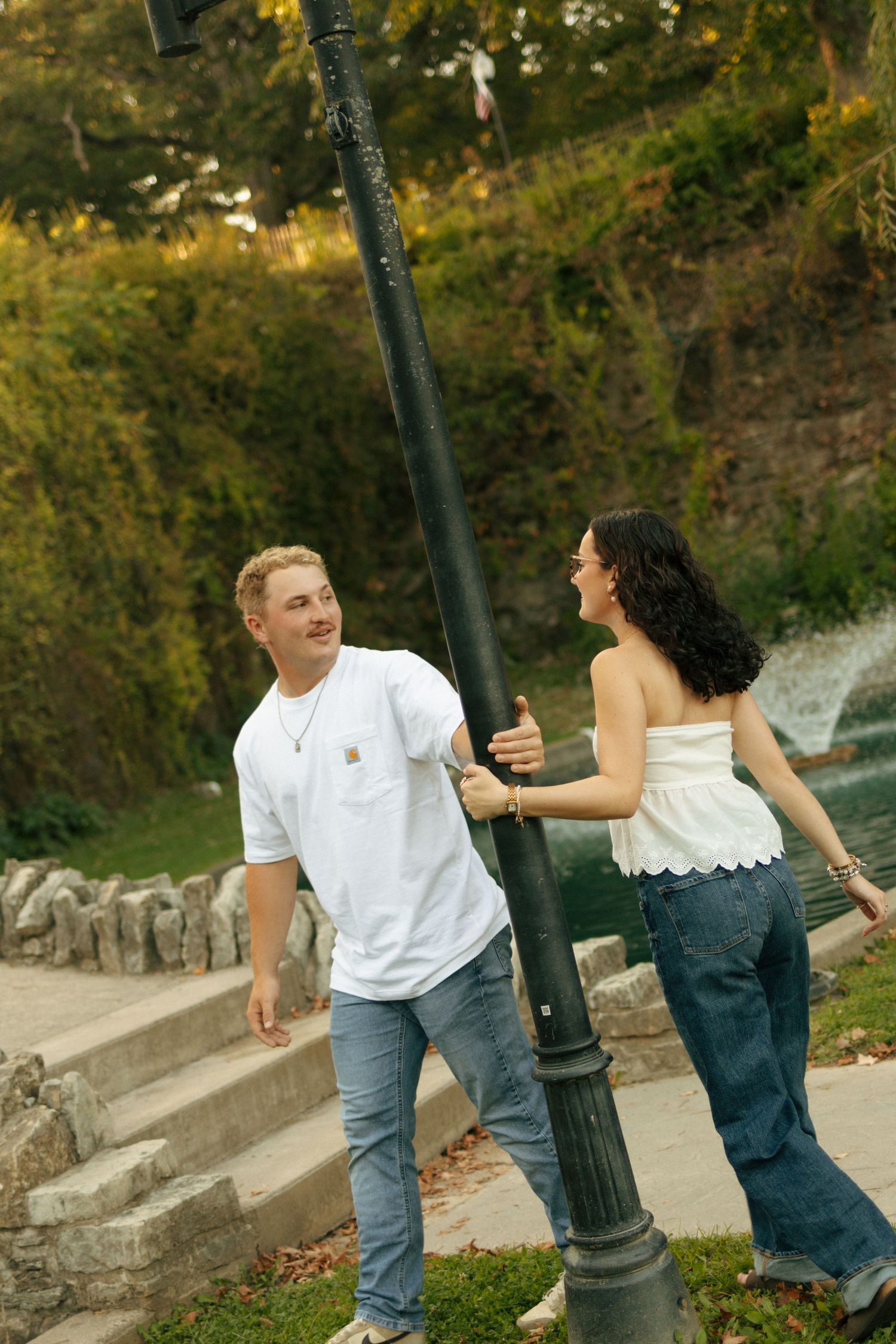 A smiling couple playfully interacts around a black lamp post in a lush green park setting.