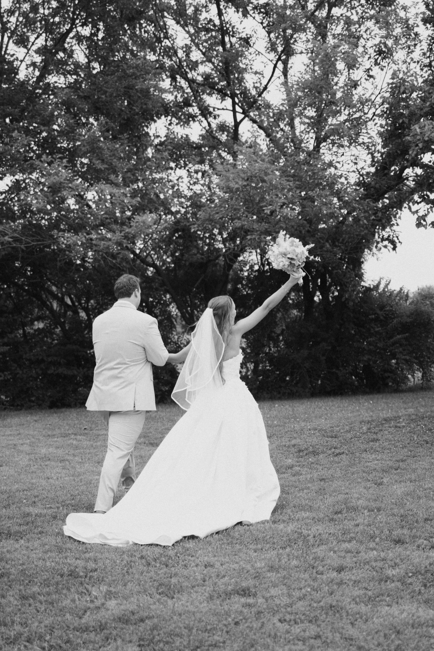 Black and white photo of bride and groom walking hand in hand on grass, bouquet raised.