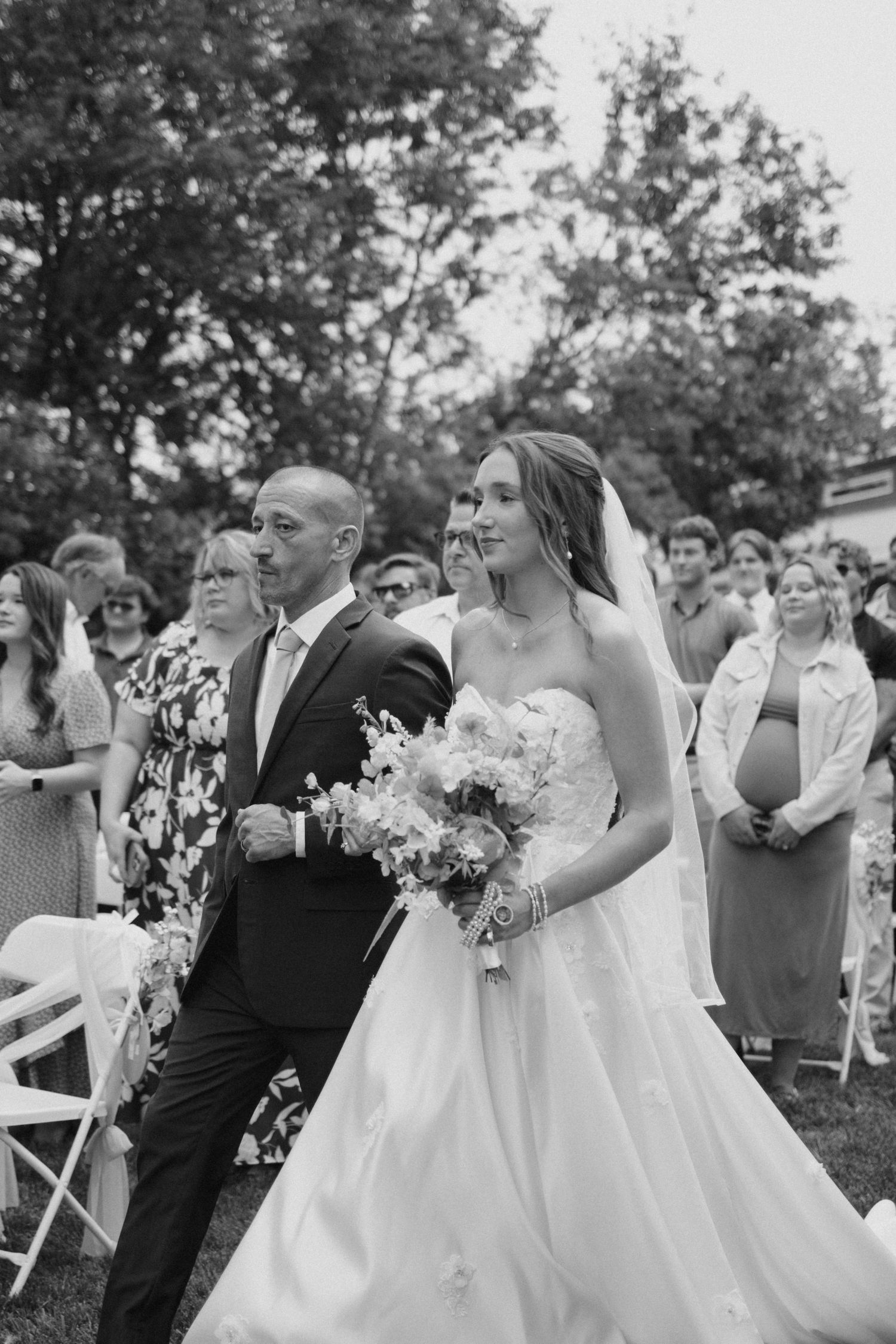 Black and white photo of a bride walking down the aisle with her escort at an outdoor wedding ceremony.