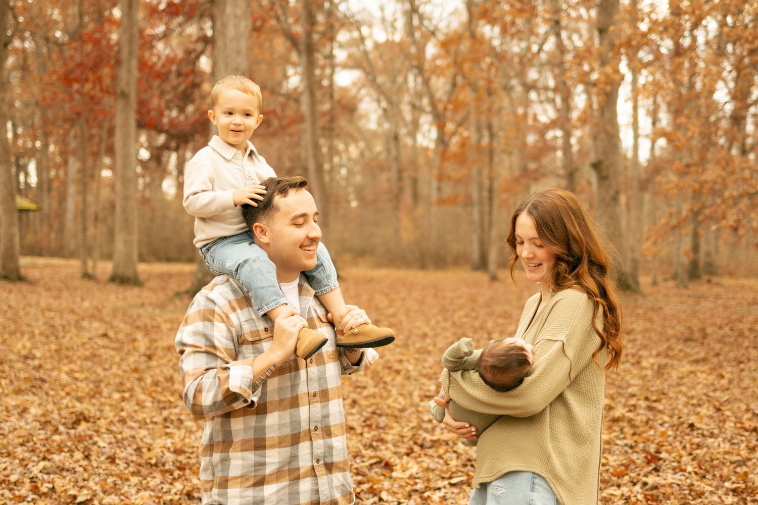 Family enjoying autumn day in forest, father carrying toddler on shoulders, mother holding baby, surrounded by fallen leaves.