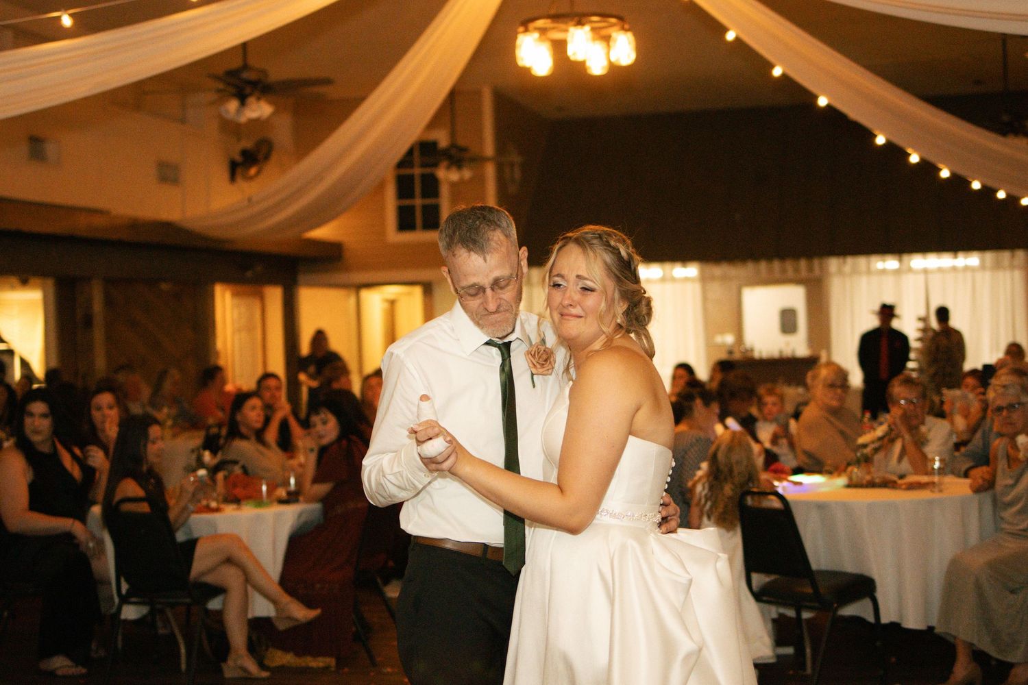 Bride and older man share father-daughter dance in decorated reception hall with string lights.