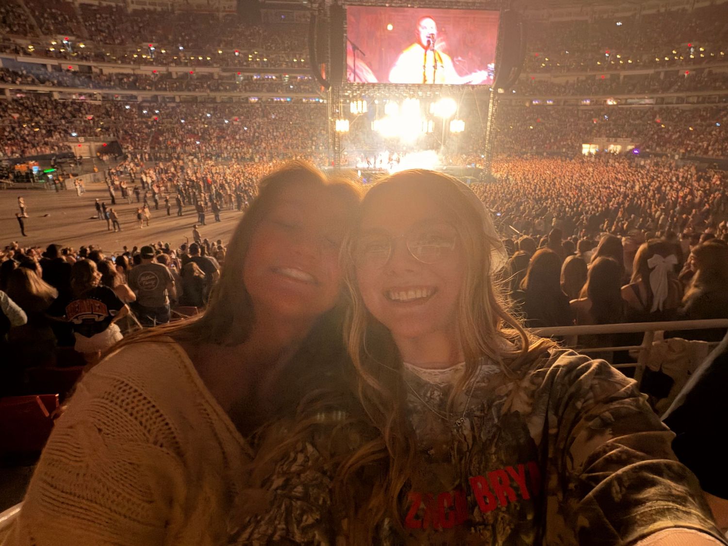 Two women smiling for a selfie at a packed indoor concert arena with bright stage lights in the background.