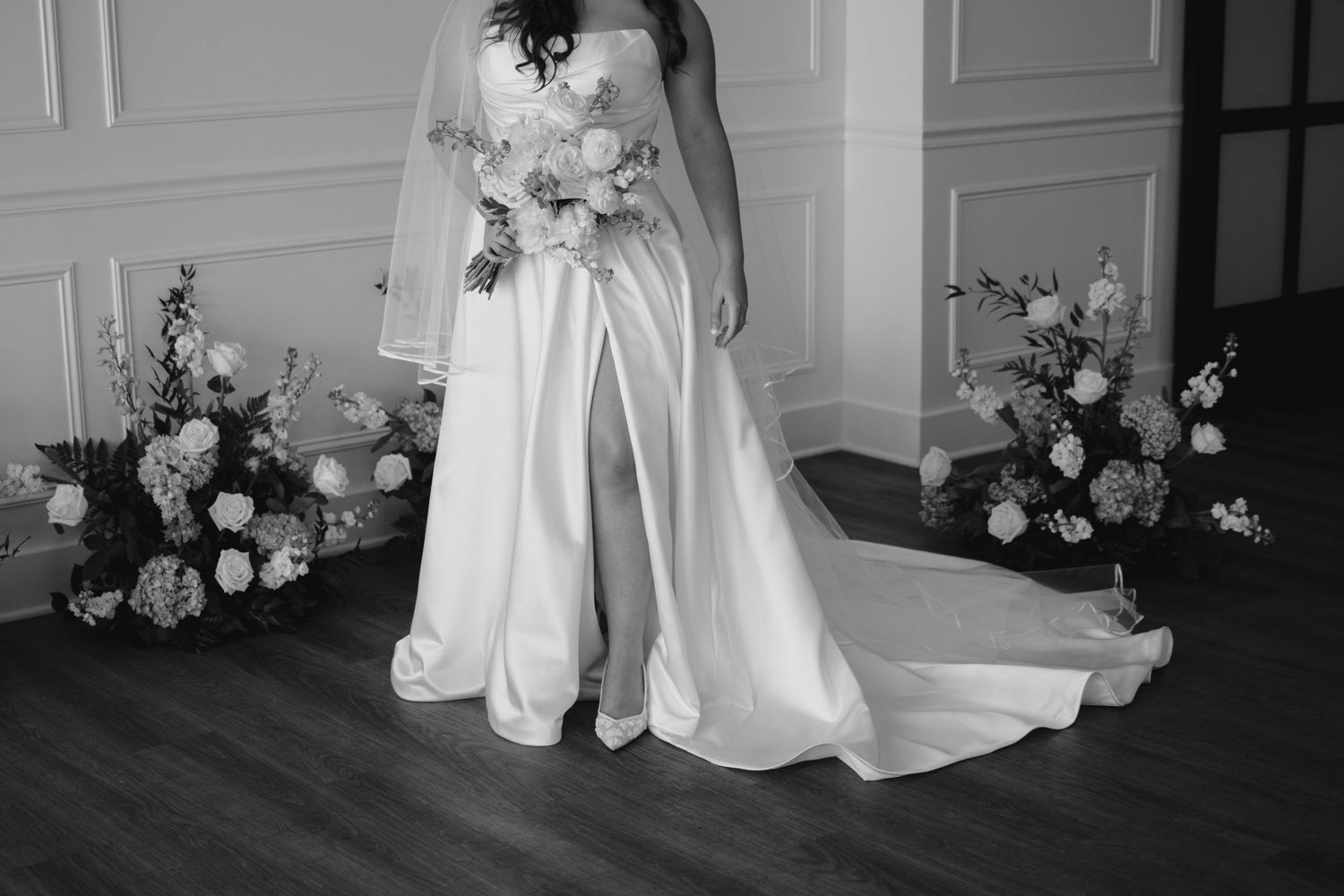 Black and white photo of a bride in a elegant gown with a slit, holding a bouquet, flanked by floral arrangements.