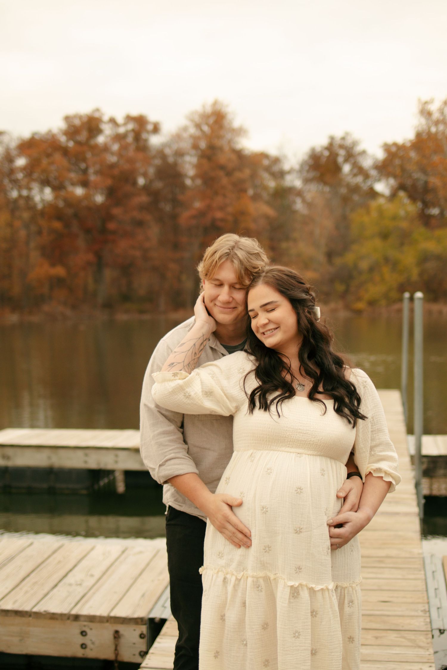 Couple sharing a tender moment on a lakeside dock in autumn, woman in white maternity dress, fall foliage background.