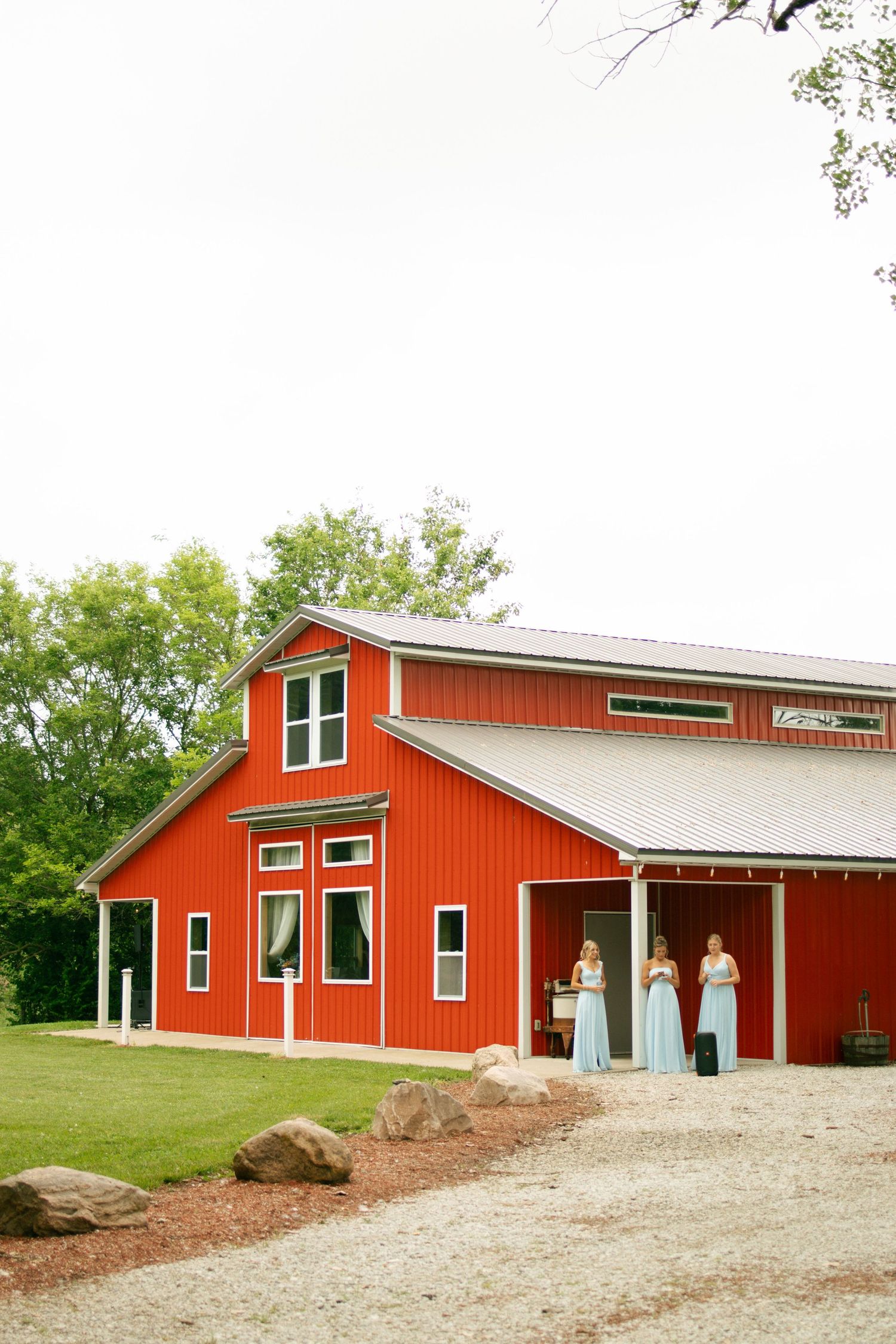 Bridesmaids in light blue dresses gather outside a rustic red barn wedding venue surrounded by lush green trees.