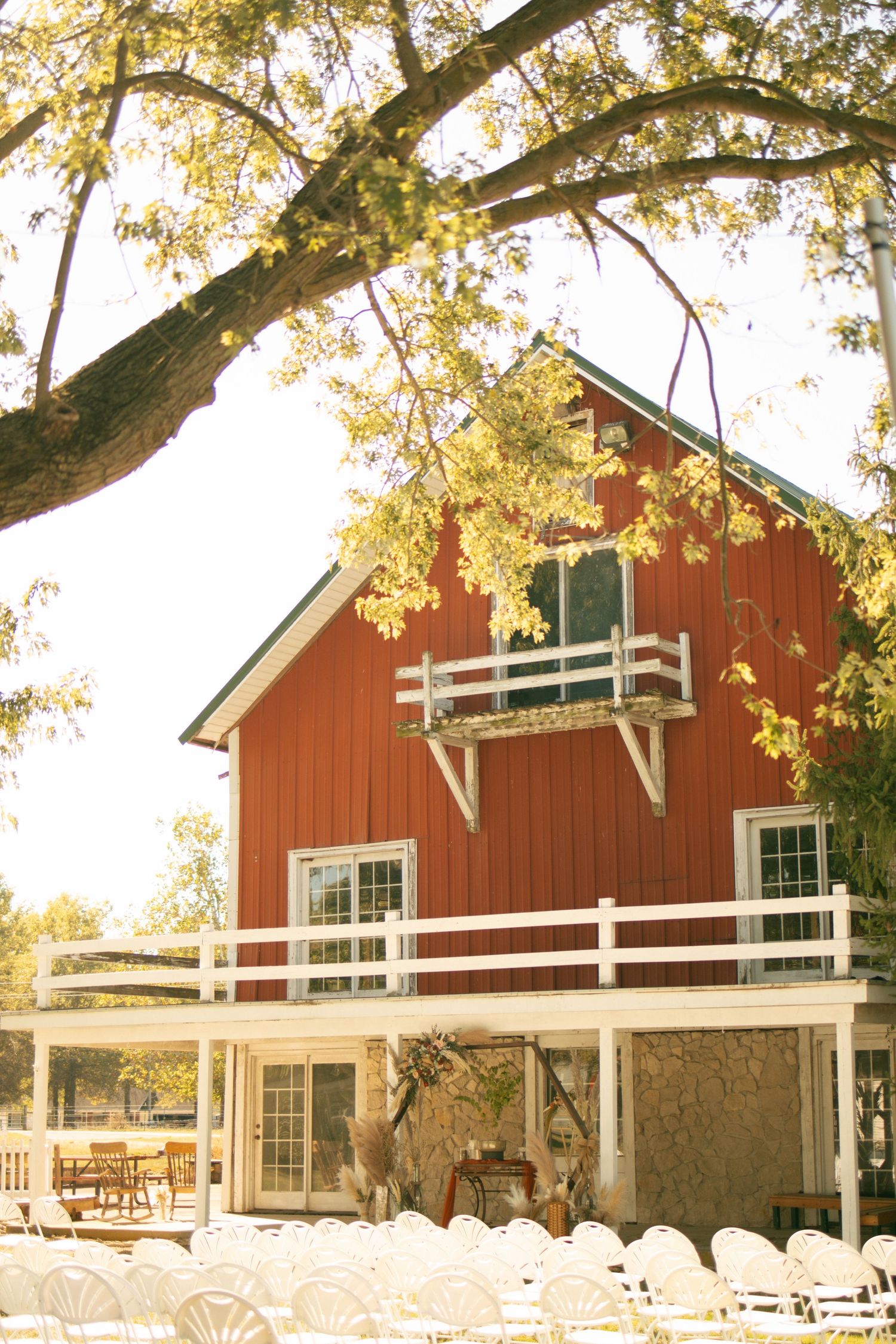 Rustic red barn wedding venue with white chairs set up outdoors under large oak trees.