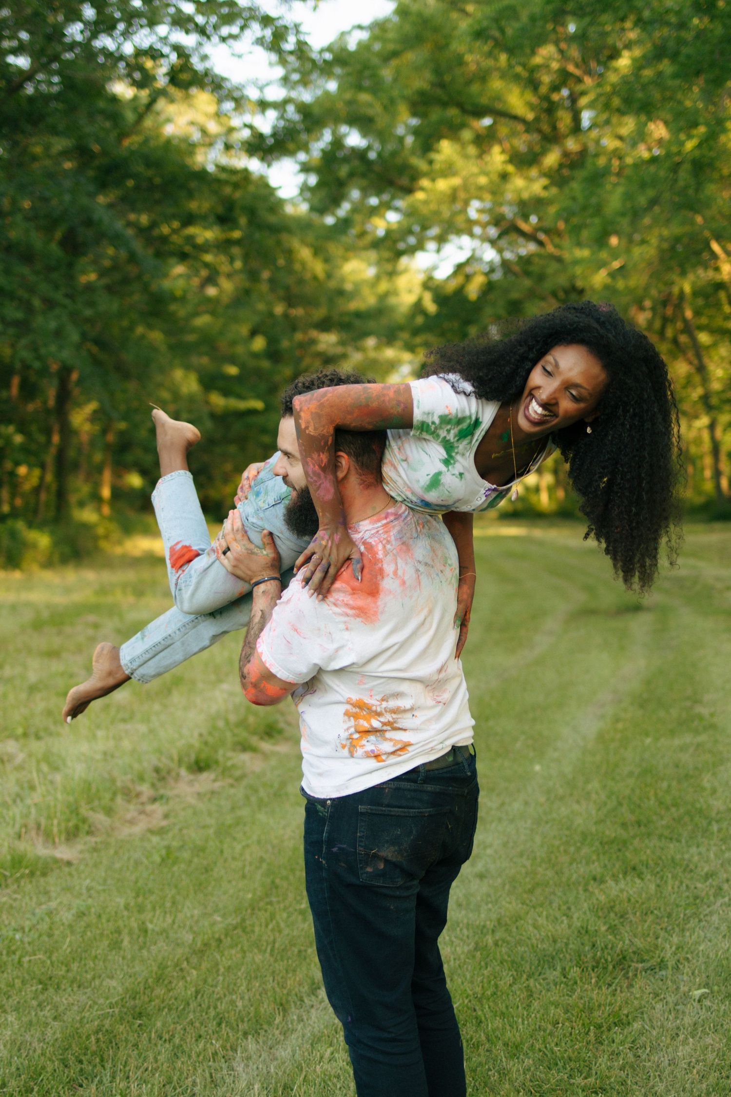Joyful man lifting laughing woman in paint-covered clothes in a green park at sunset.