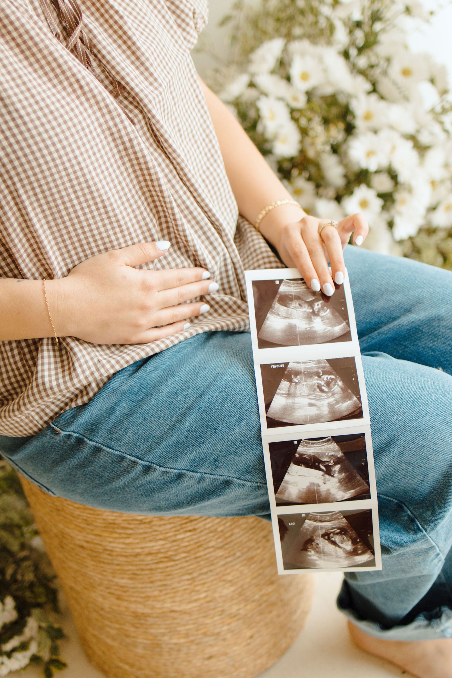 Pregnant woman in jeans holding ultrasound photos while sitting near white flowers, gently touching her baby bump.