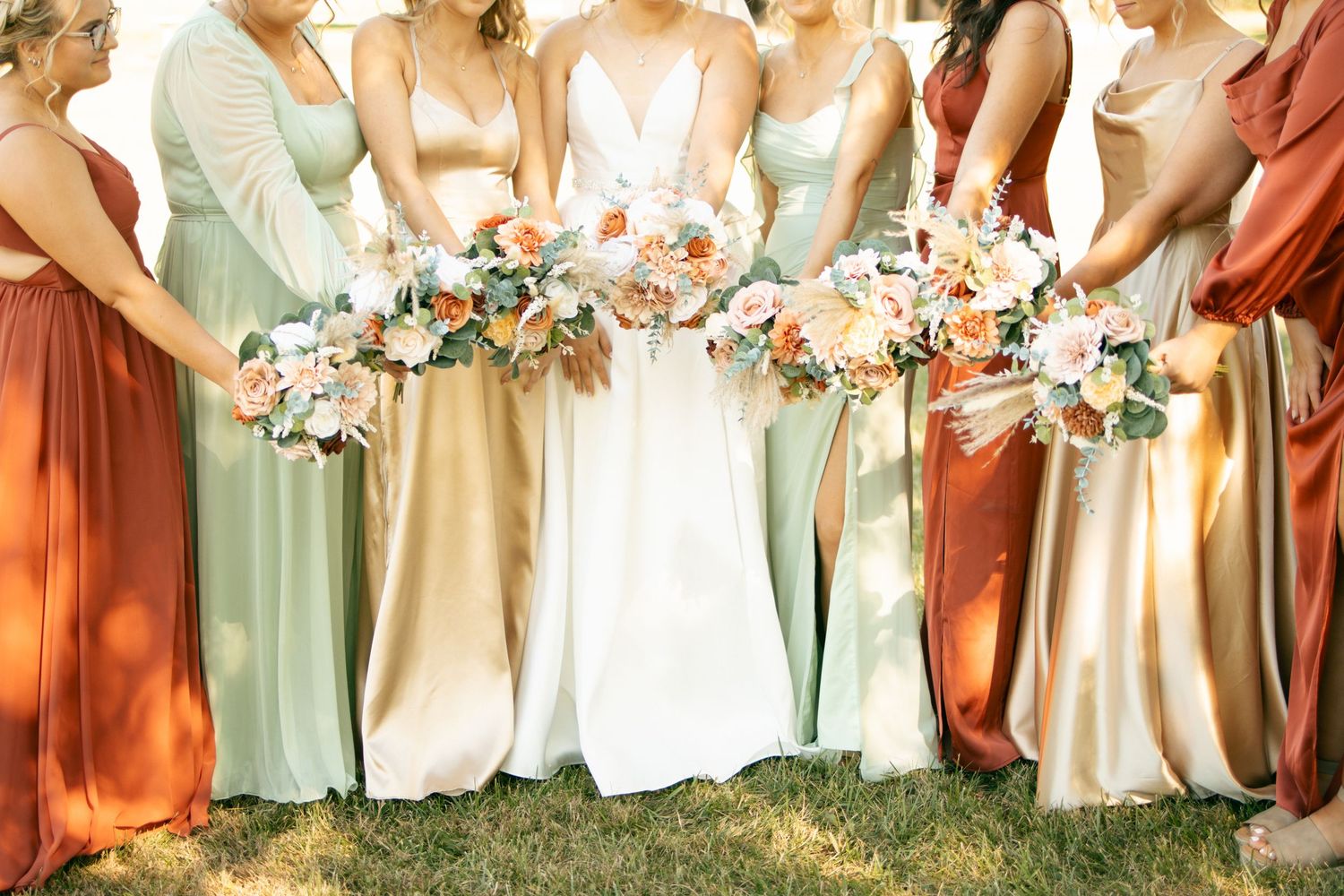 Bride and bridesmaids in sage green, gold, and rust dresses holding peach and white floral bouquets.