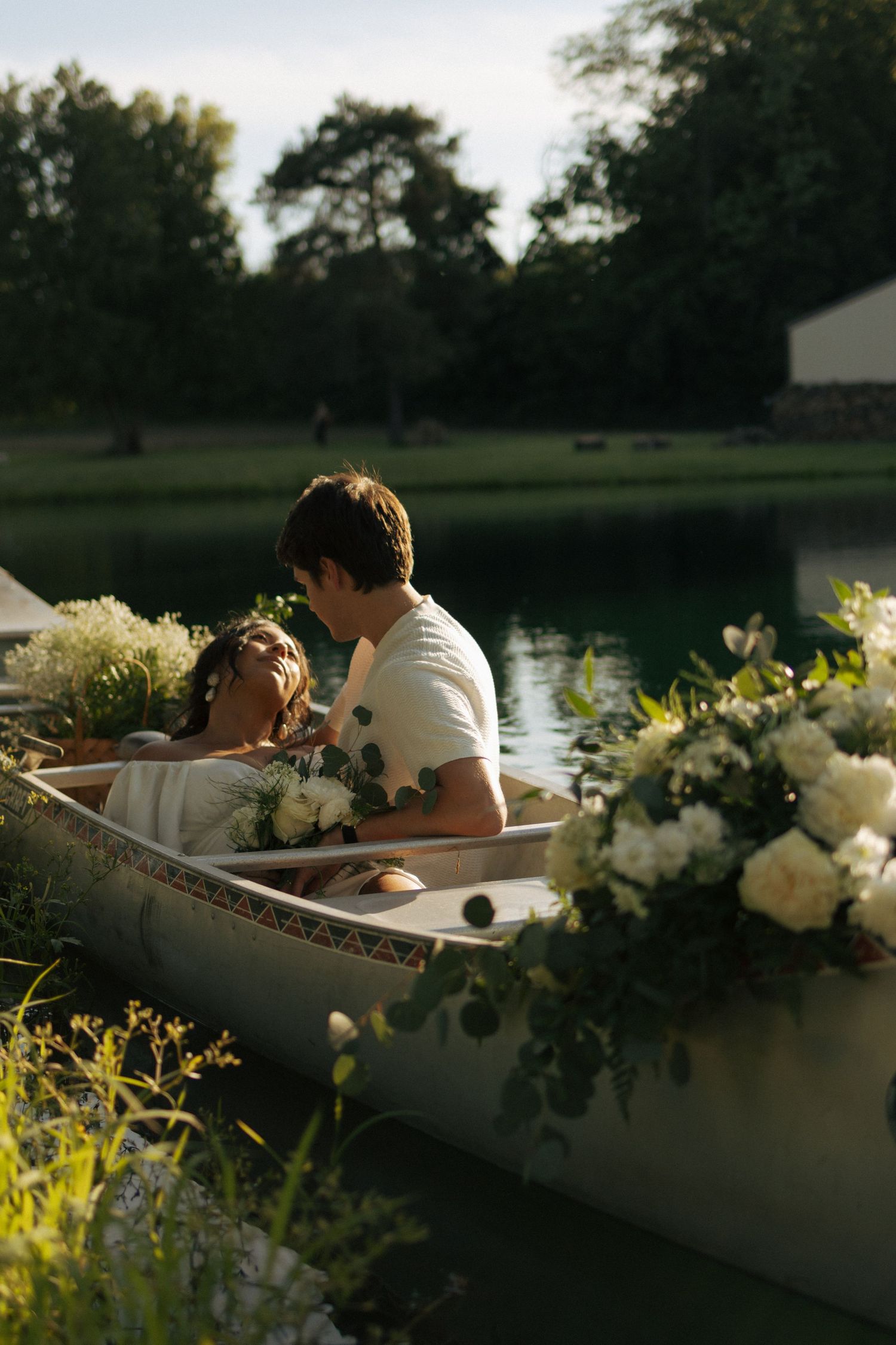 A couple sits in a flower-adorned canoe on a serene lake surrounded by lush greenery.