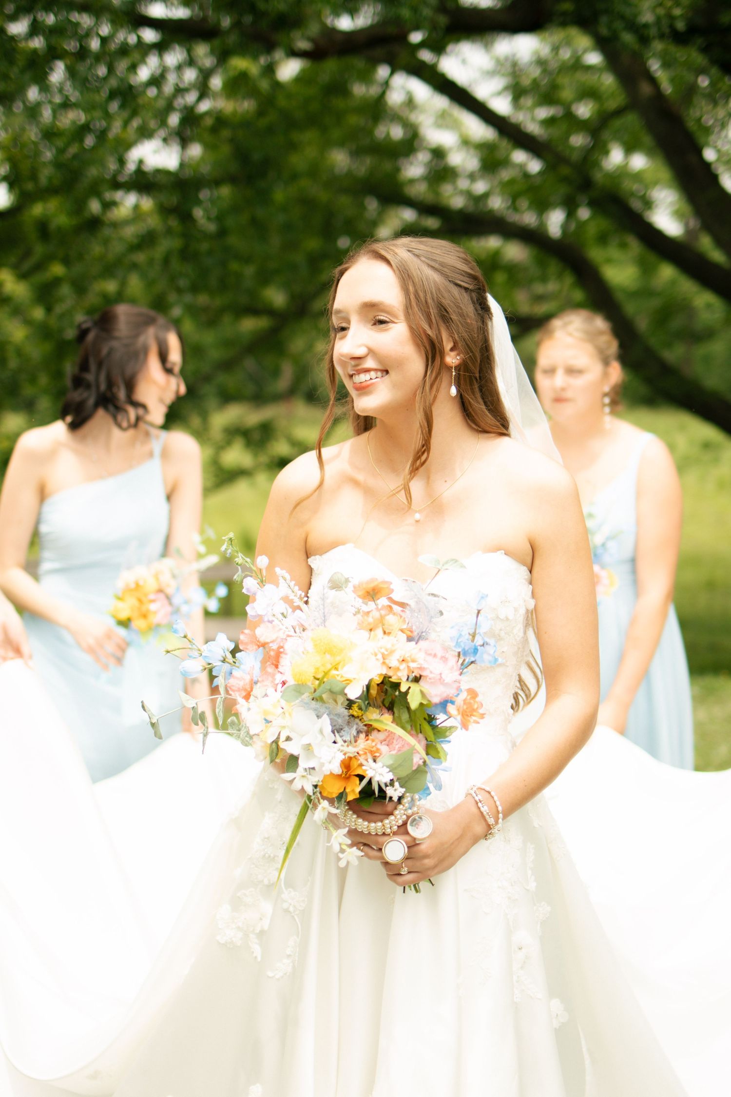 Smiling bride in strapless white gown holds colorful wildflower bouquet with bridesmaids in light blue dresses behind her.