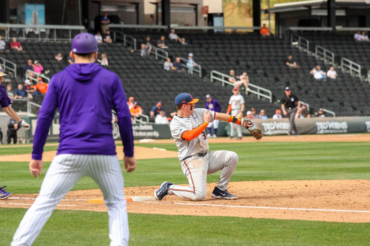A baseball player kneels in batting stance at home plate while a player in purple stands nearby during a game.