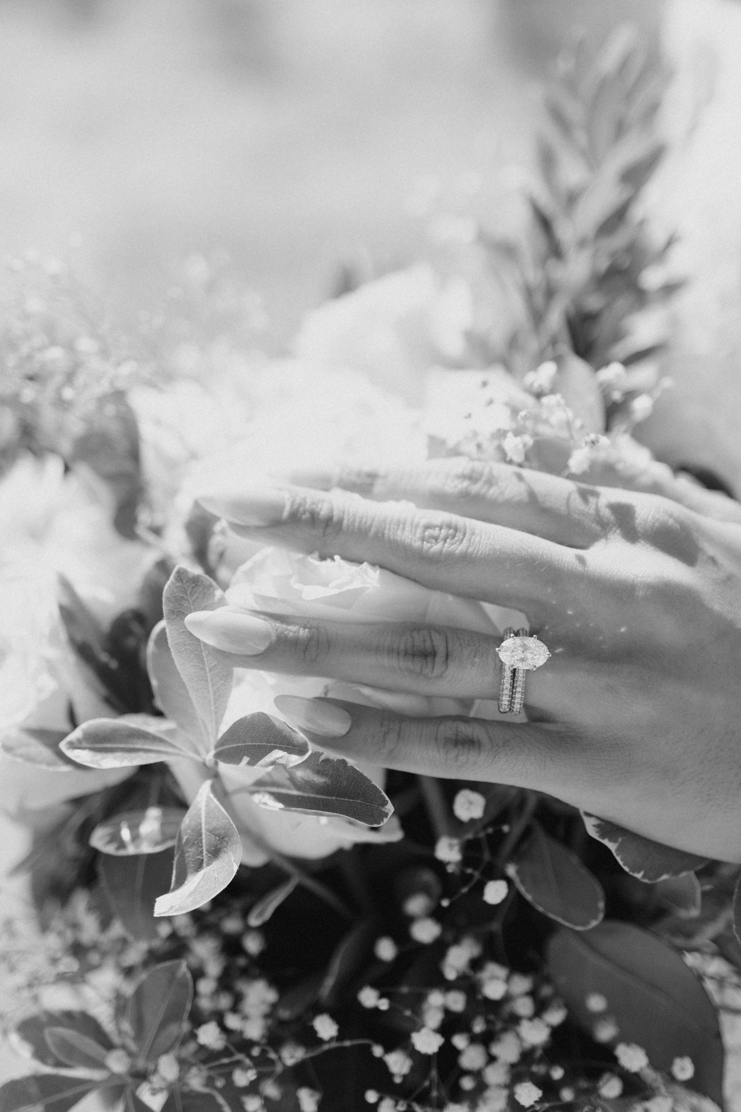 Elegant engagement ring shown in black and white with floral bouquet in soft focus background.