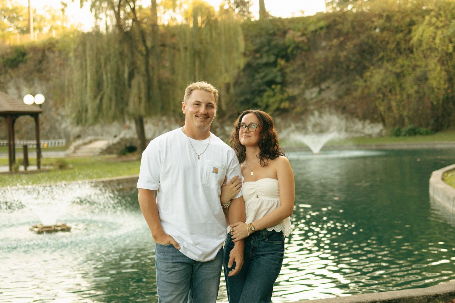 A couple embraces during their engagement photo session at a scenic lake with a fountain at sunset.