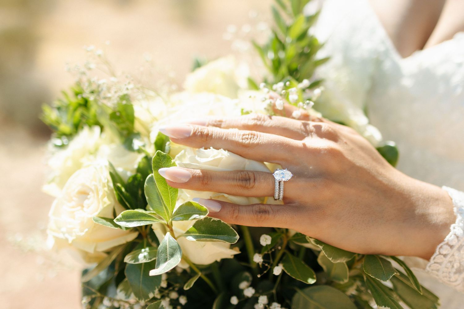 Hand with engagement ring resting on white rose and greenery wedding bouquet in soft natural lighting.