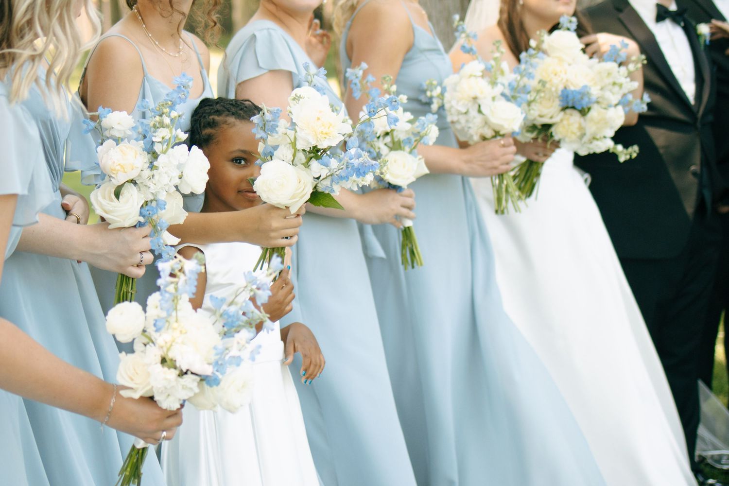 A young flower girl in white peeks between bridesmaids in light blue gowns holding white and blue floral bouquets.