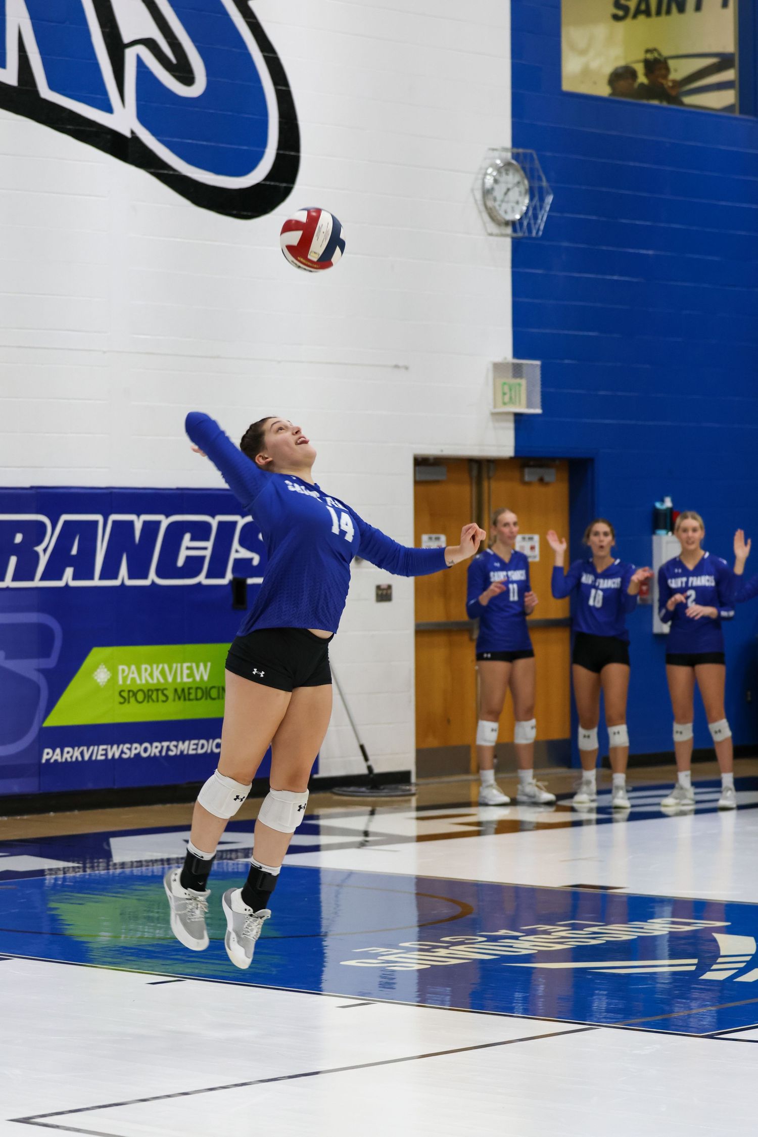 A volleyball player in blue uniform jumps to serve the ball during a match while teammates watch from the sideline.