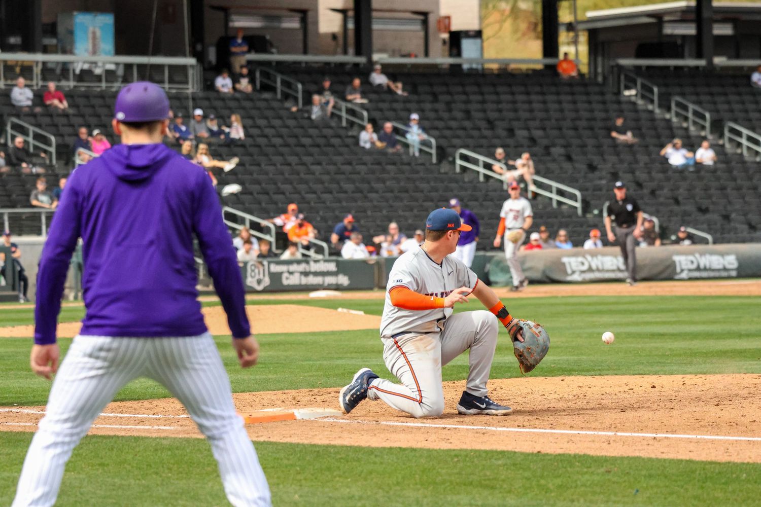 Baseball players in action during a game with one player fielding a ground ball while a coach watches from nearby.