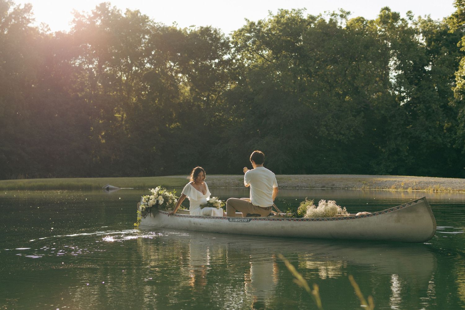 Bride and groom paddling a canoe across a serene lake at golden hour, surrounded by lush green trees.